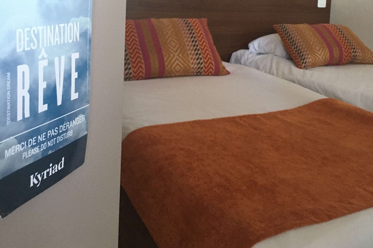 A hotel room with two beds, a world map on the wall, a lamp, and a sign with the hotel name 'Kyriad'.