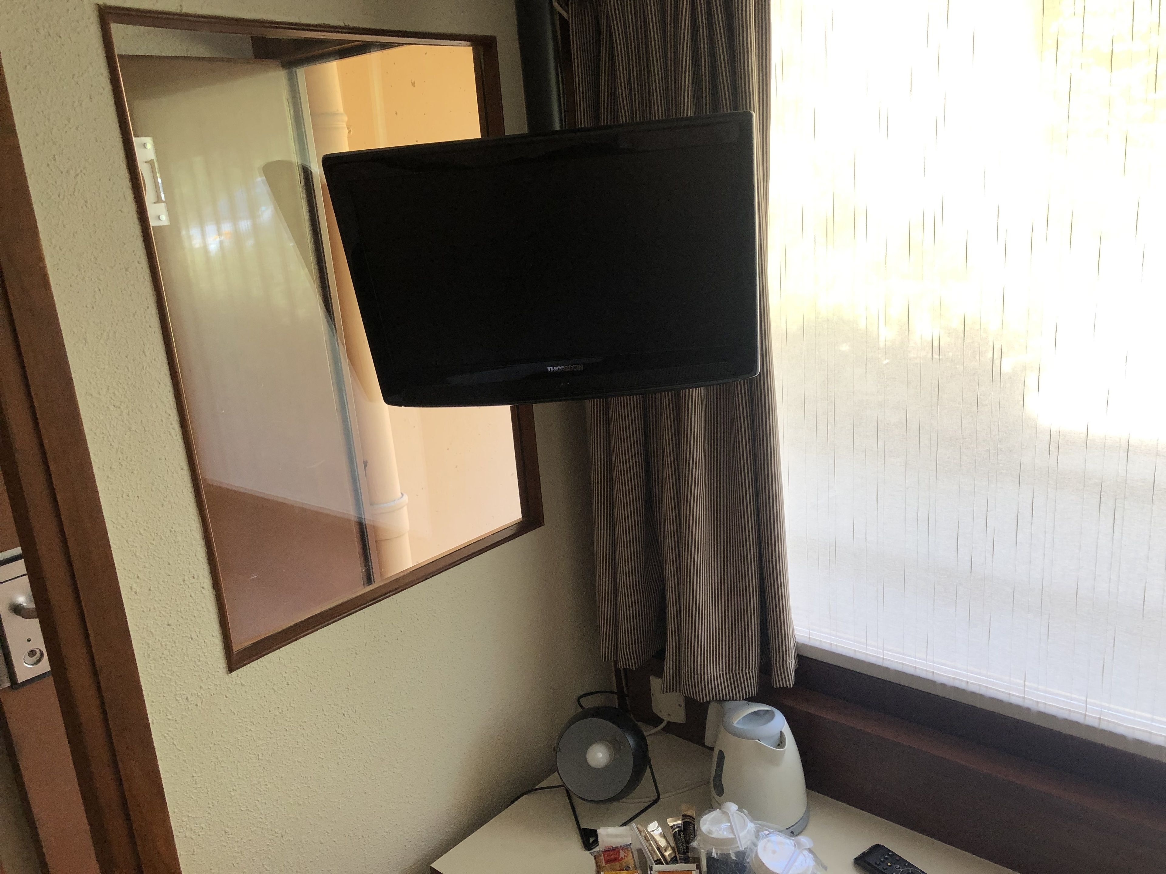 Ein Hotelzimmer mit Flachbildfernseher an der Wand, einem Fenster mit Jalousien, einem kleinen Tisch mit Wasserkocher, einer Fernbedienung und einem Spiegel.