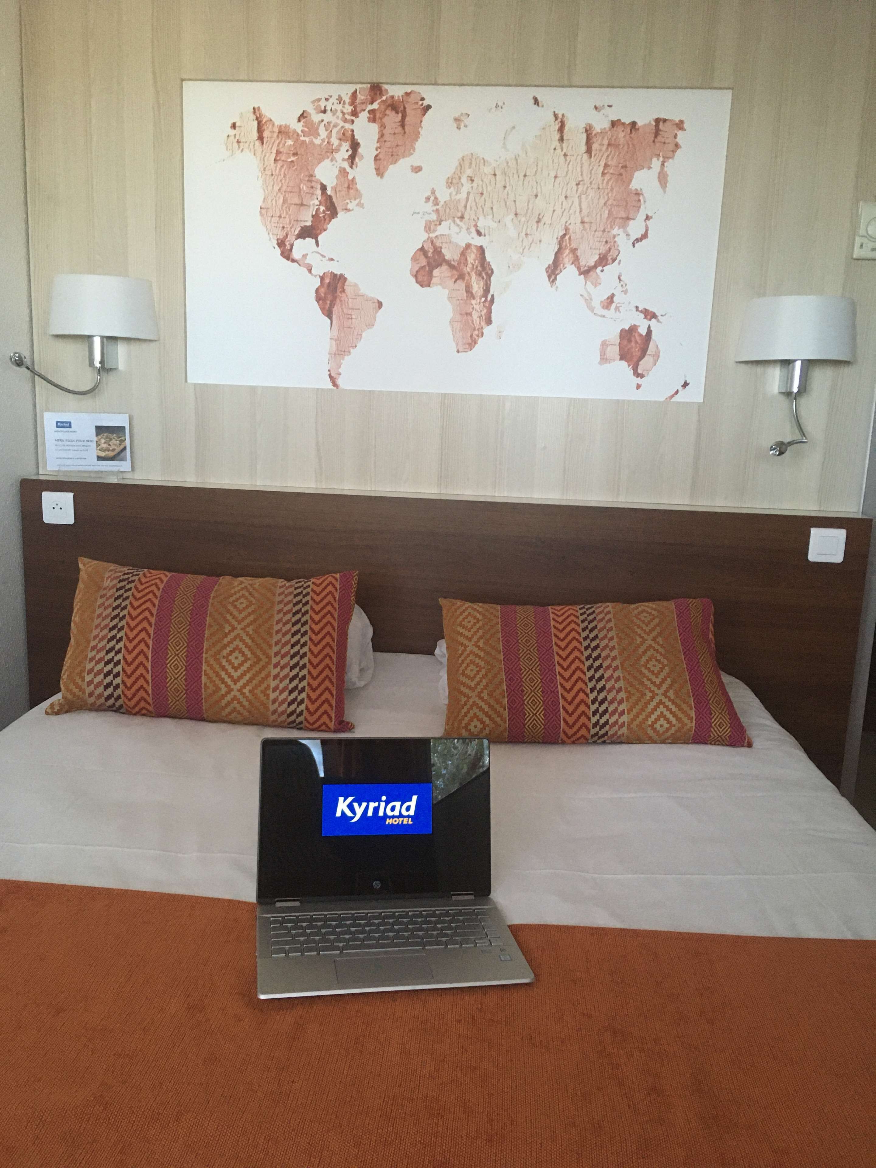 Quarto de hotel com uma cama, duas almofadas estampadas, um laptop na cama, um mapa-múndi na parede e duas luminárias de parede.