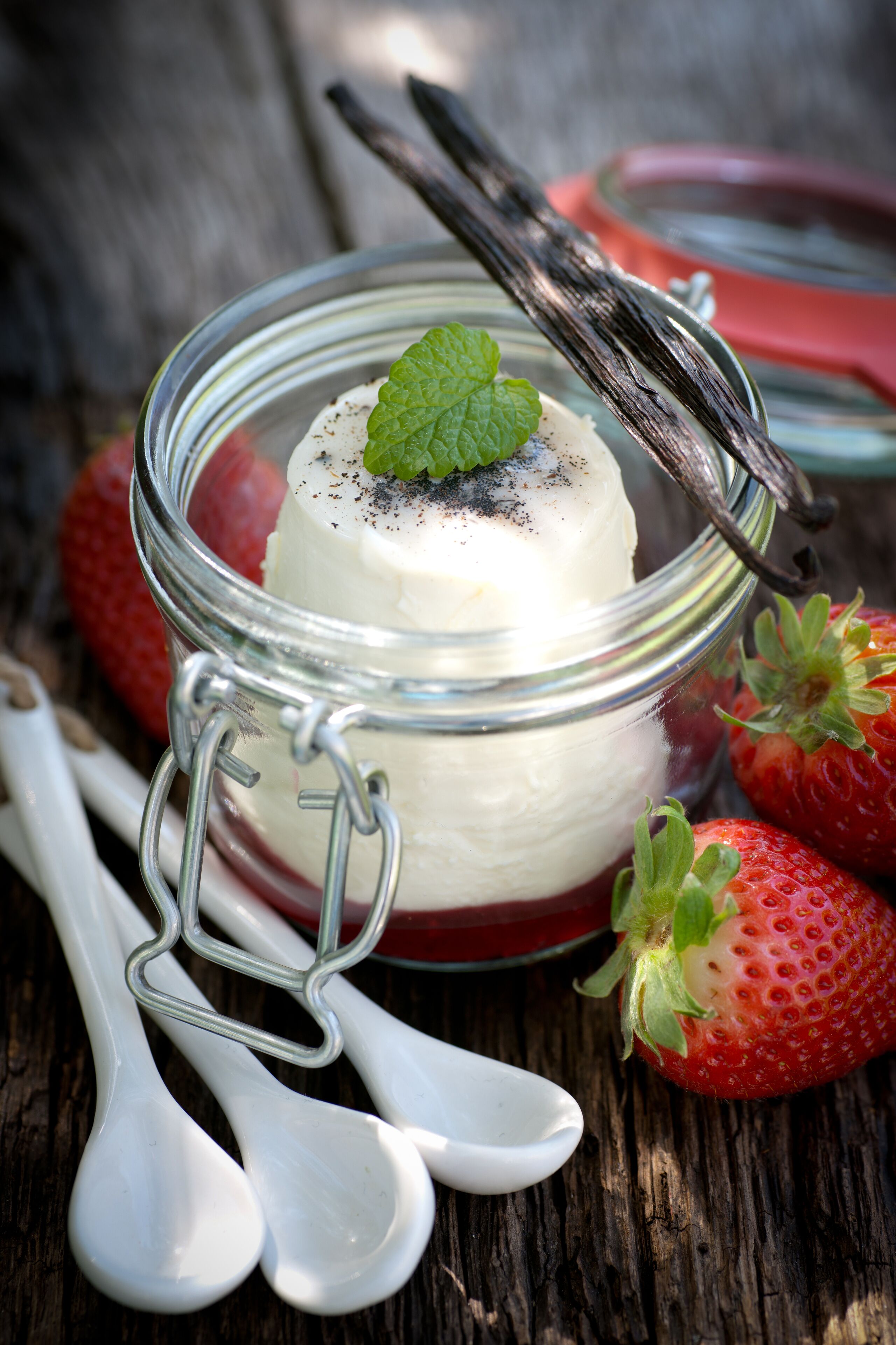 Postre en un frasco de vidrio con cierre metálico, acompañado de fresas y cucharas sobre una superficie de madera.