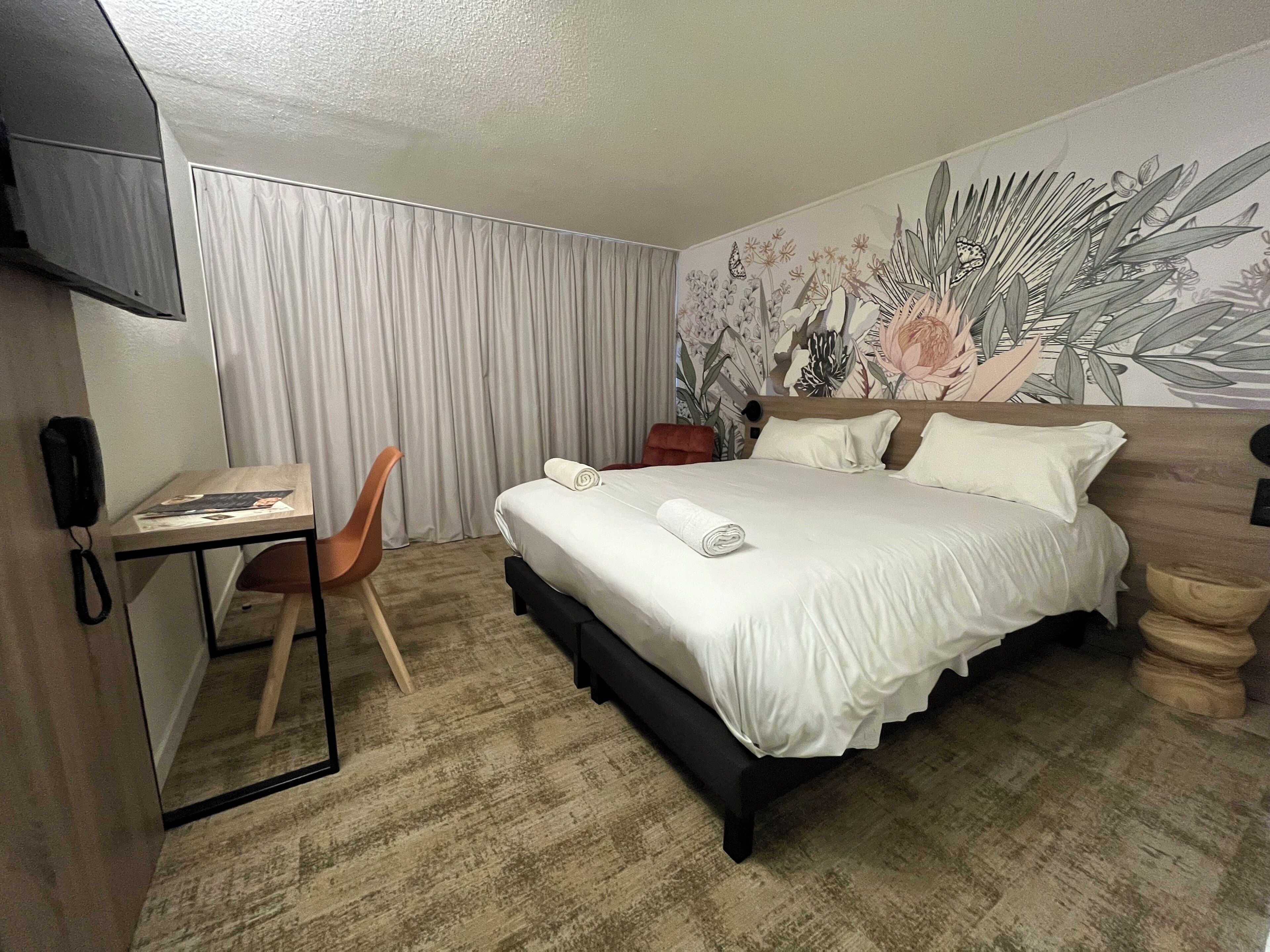 A hotel room with a bed, a desk, a chair, a telephone, and a wall with a floral mural.