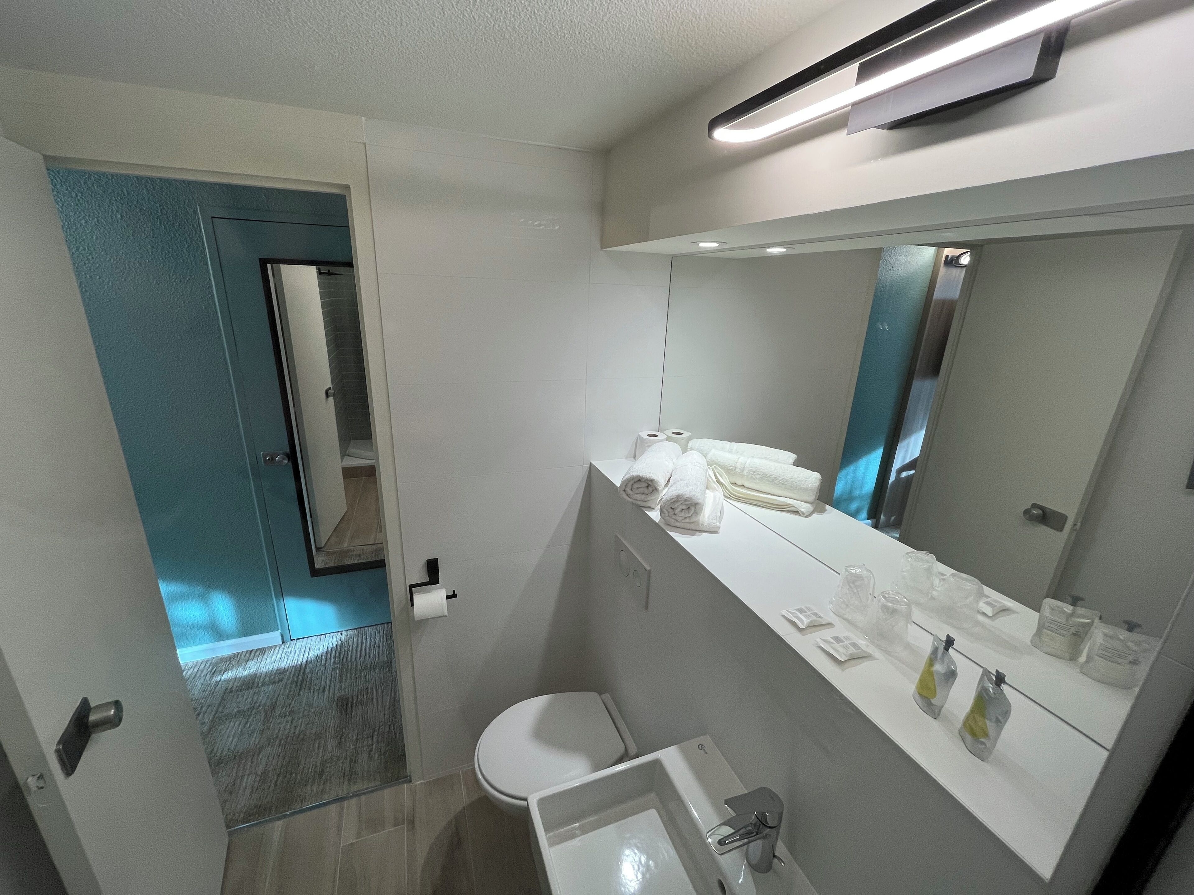 Un baño moderno con inodoro, lavamanos y toallero. Se ve una zona de ducha a través de una puerta abierta.