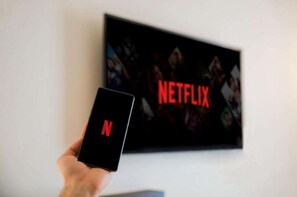 Une main tenant un smartphone sur lequel figure le logo de Netflix, avec un téléviseur affichant également le logo de Netflix en arrière-plan.