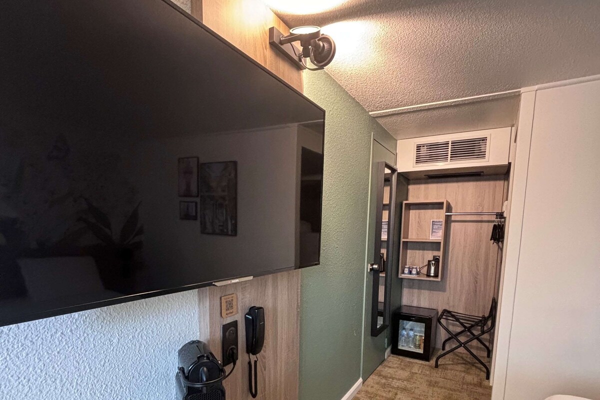 Habitación de hotel con un televisor de pantalla plana grande montado en la pared, un escritorio de madera con un teléfono y una cafetera pequeña, y una cama con una colcha blanca.