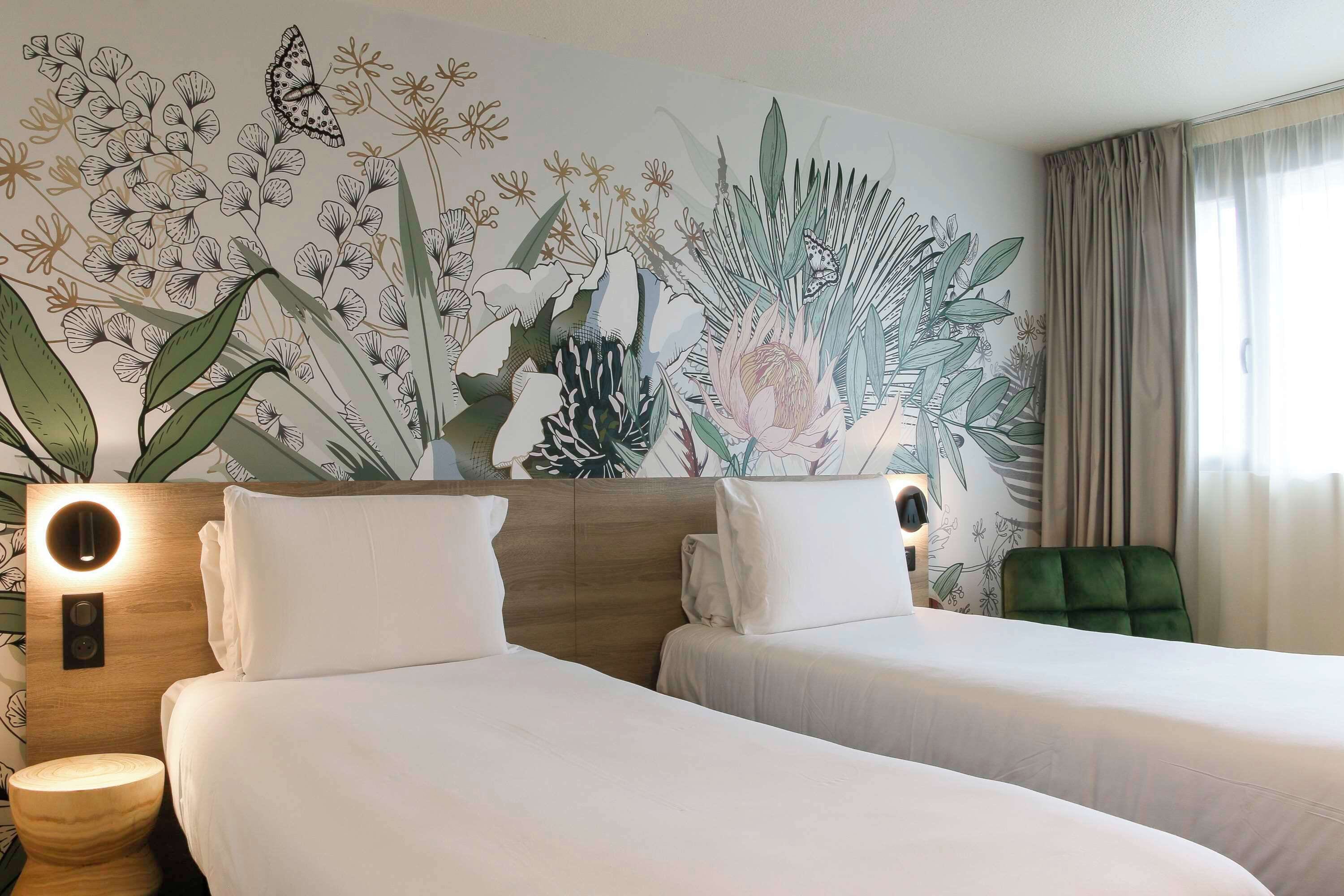 Une chambre d’hôtel avec deux lits, un fauteuil vert et une fresque murale représentant des fleurs et des plantes.