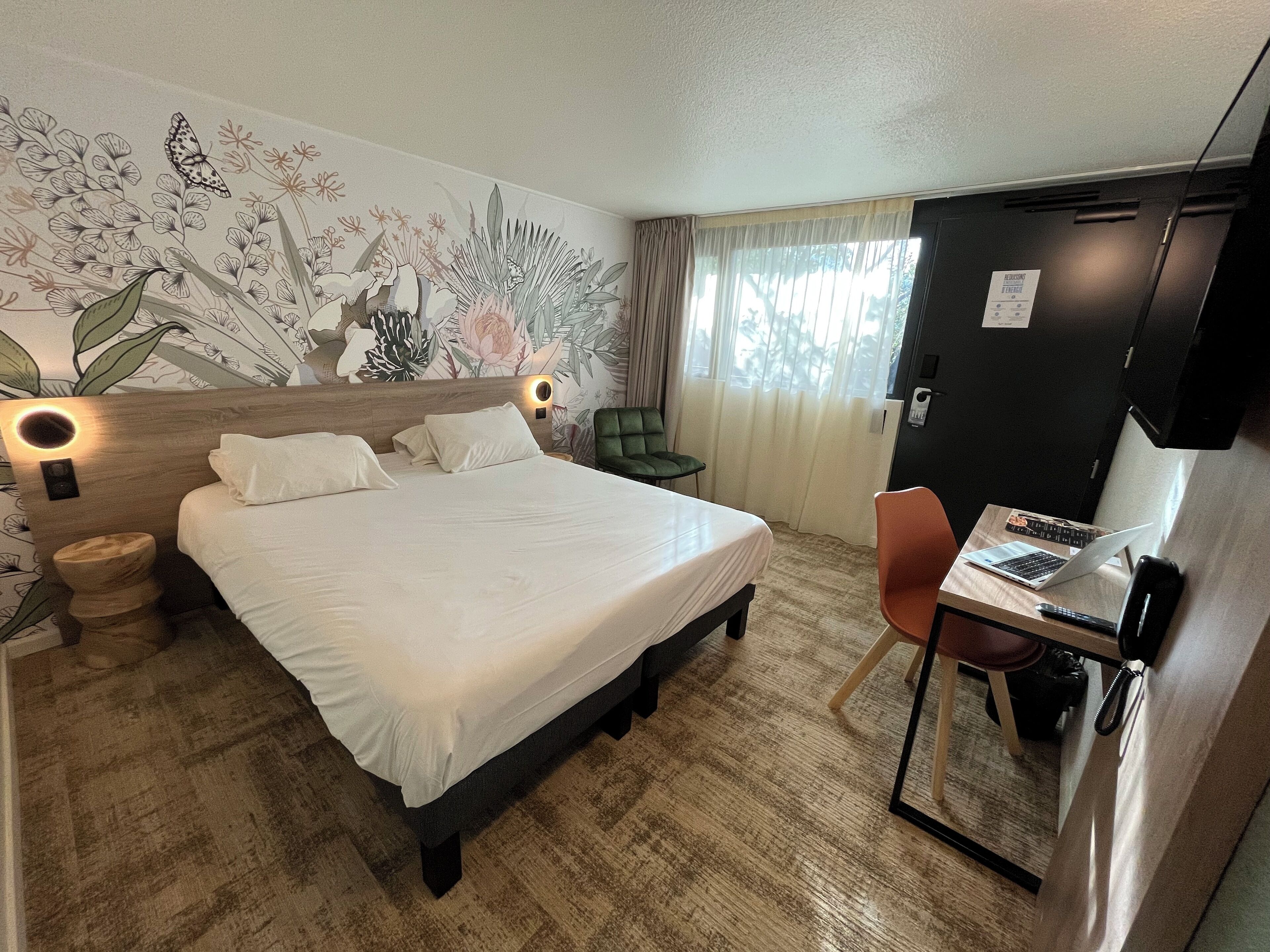 A hotel room with a large bed, a desk with a laptop, a green chair, a wooden nightstand, and a wall with a floral mural.
