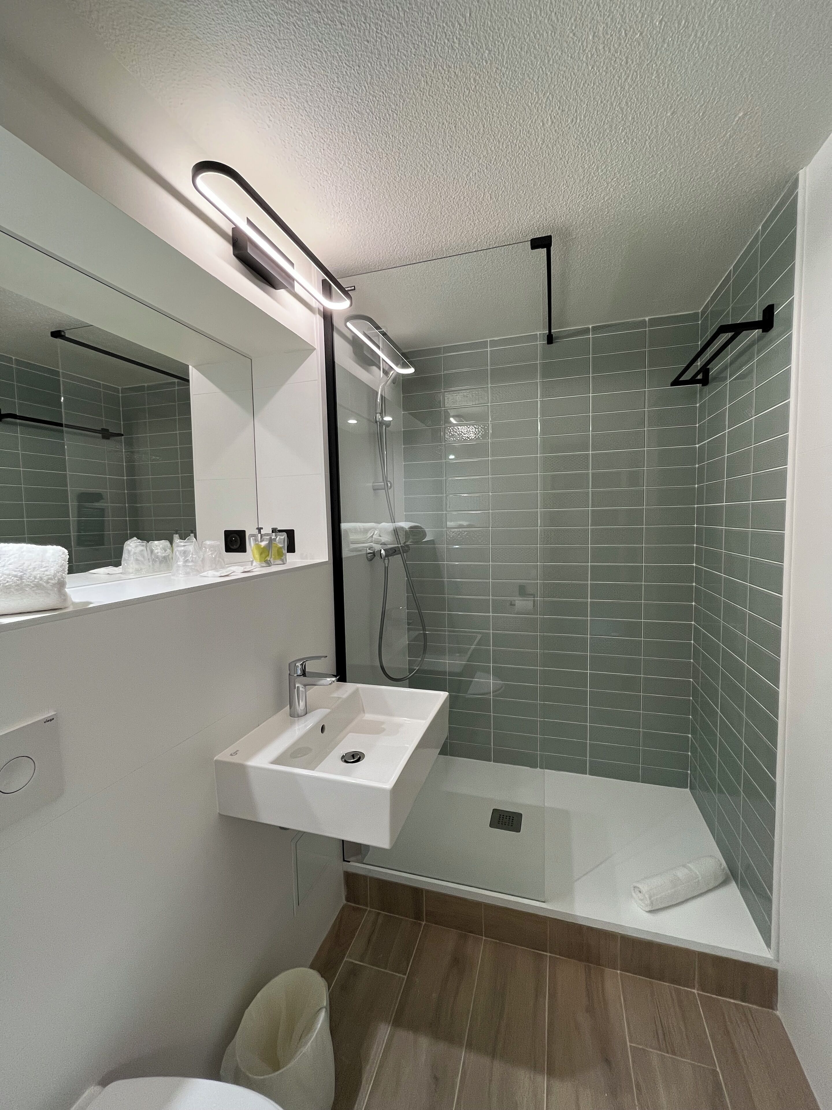 Un baño moderno con ducha envuelta en vidrio, lavamanos blanco e iluminación integrada en el espejo.