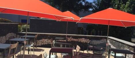 An outdoor seating area with tables and chairs under red umbrellas, surrounded by stone walls and trees.
