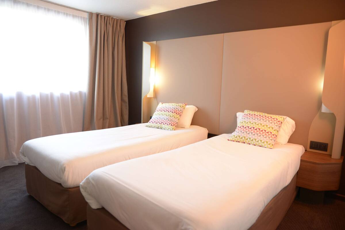 A hotel room with two beds, each with a white bedspread and patterned pillows, a large window with curtains, and a wall-mounted lamp.