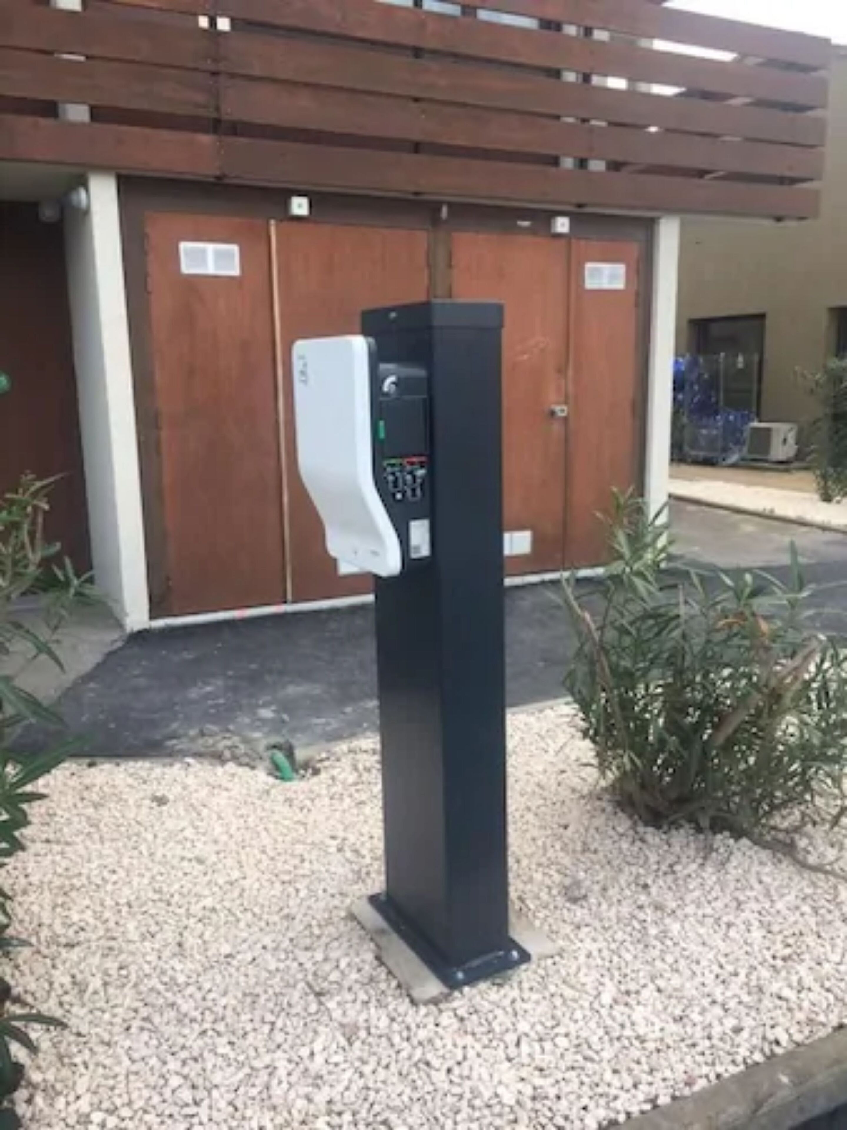 A black vending machine with a white dispenser in front of a building with a wooden facade and a gravel ground.