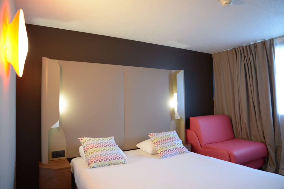 A hotel room with a bed, two pillows, a red chair, a nightstand, and a wall-mounted lamp.