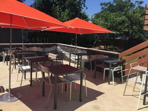 Ein Außenbereich mit Terrasse, Tischen, Stühlen und roten Sonnenschirmen, mit einer Steinwand im Hintergrund.
