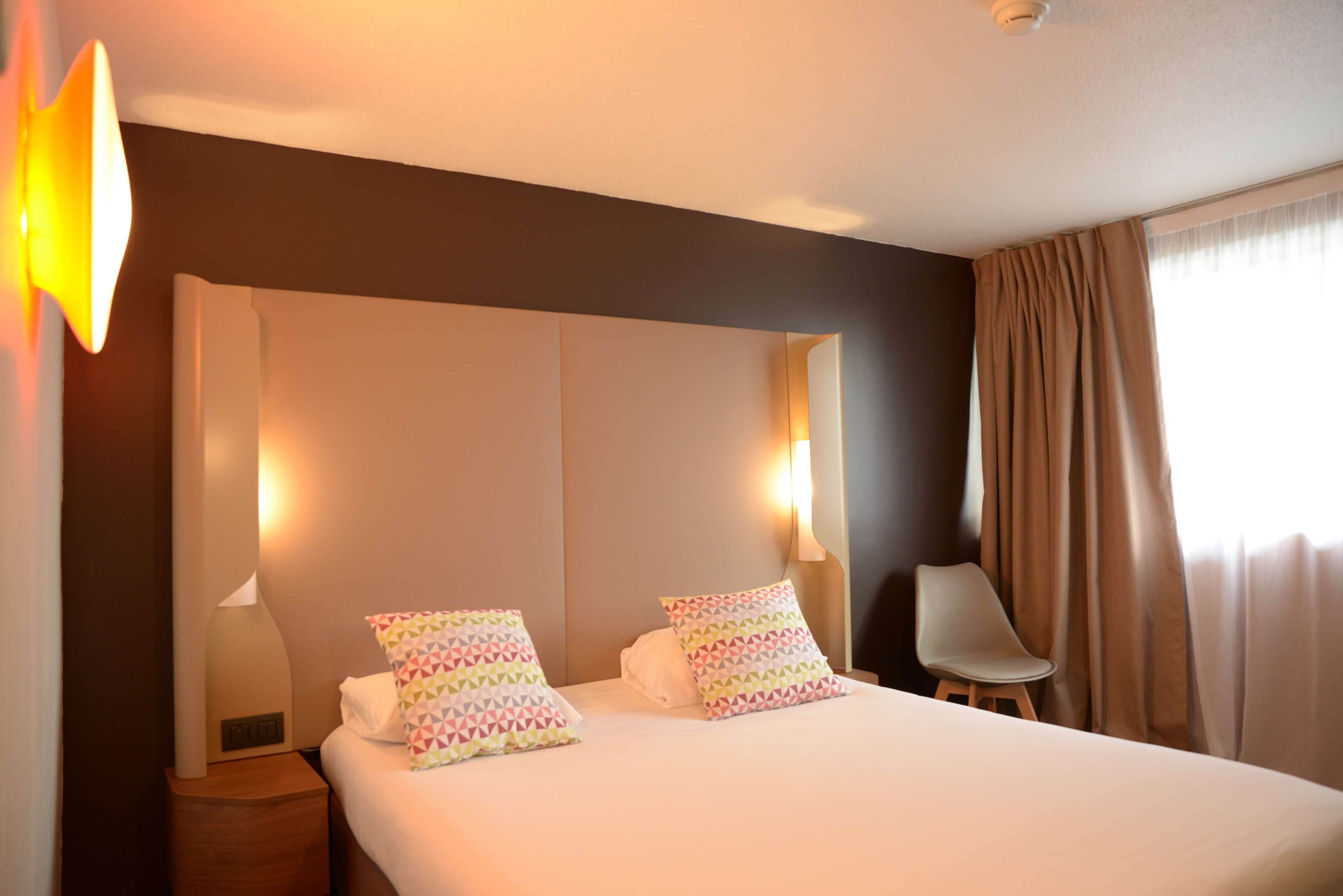 Una habitación de hotel con una cama grande, dos almohadas, una mesita de noche, una silla y una ventana con cortinas.