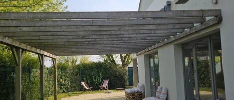 Un patio techado con mobiliario exterior, una pérgola y vistas a un edificio blanco.