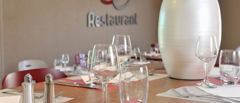 Una mesa de comedor dispuesta con copas de vino, cubiertos y saleros y pimenteros, con una planta en maceta y un letrero que dice "Le Restaurant" al fondo.