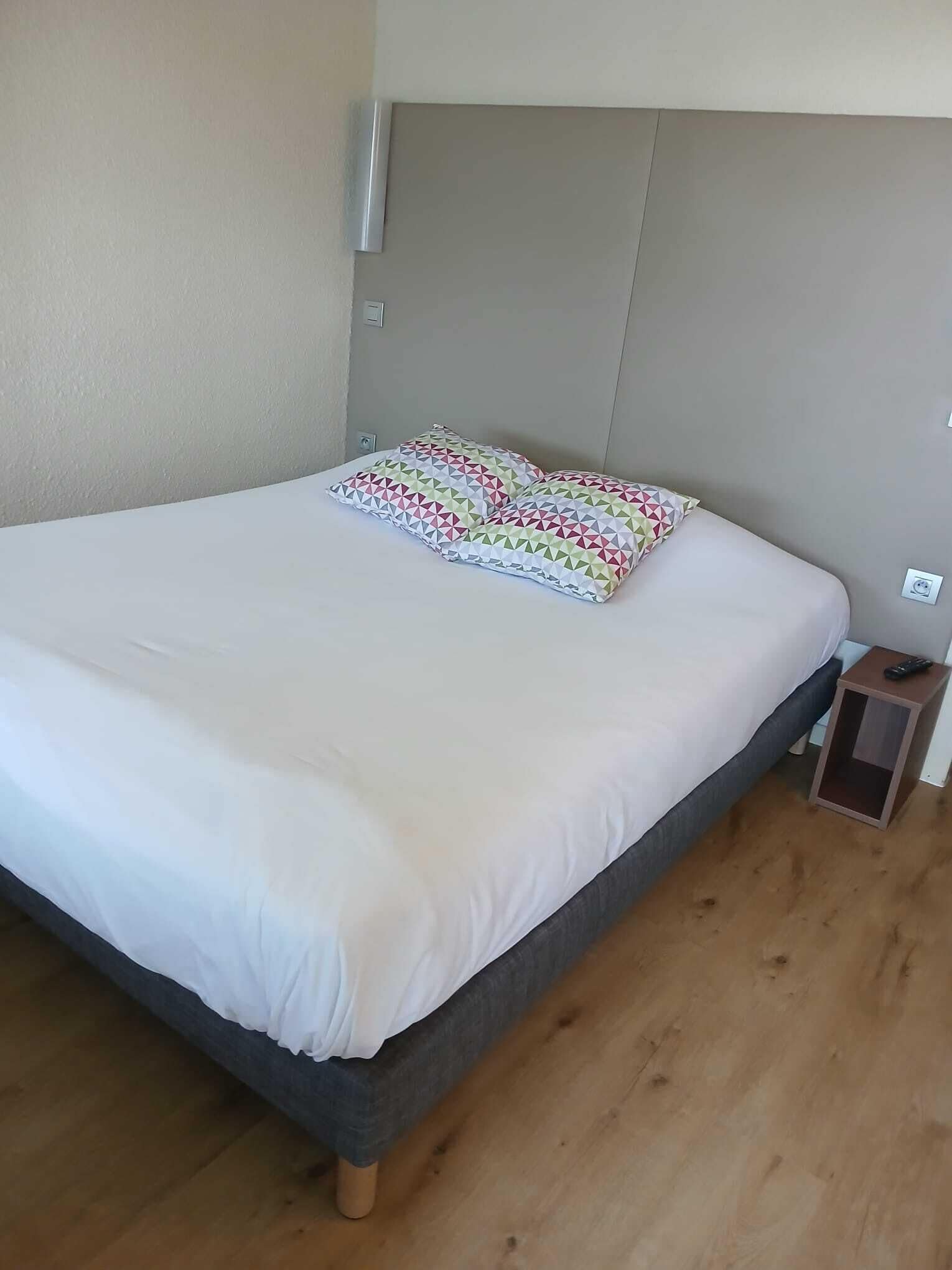 A neatly made bed with a white duvet and patterned pillows, a small wooden nightstand with a remote control, and a plain wall with a light switch.