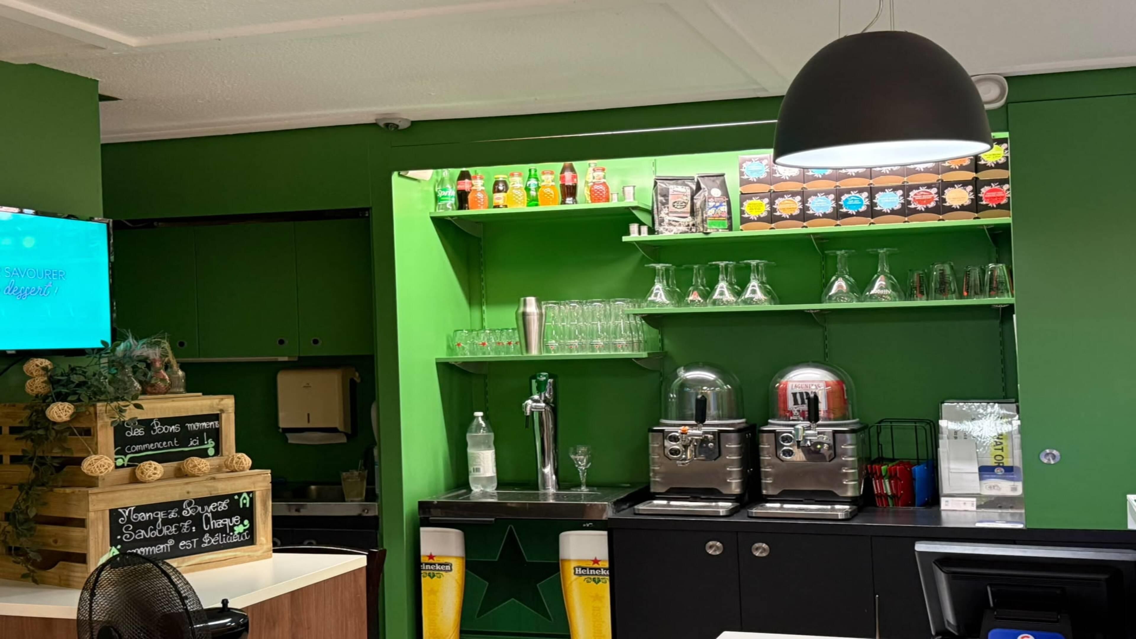 A bar area with a green wall, shelves with bottles, glasses, and a tap. There are two beer taps and a coffee machine.