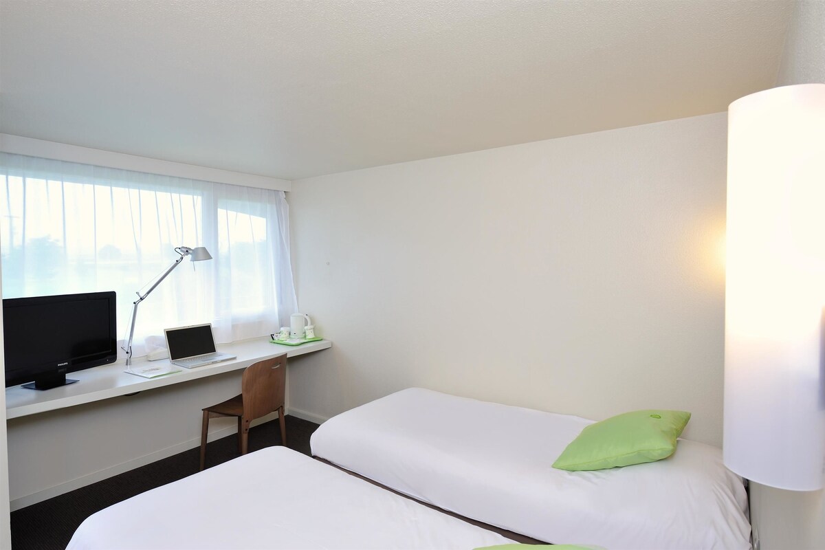 A hotel room with a desk, two beds, a chair, a computer, and a lamp.