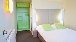 Una habitación de hotel con una cama, un armario verde, un radiador y una lámpara fijada a la pared.