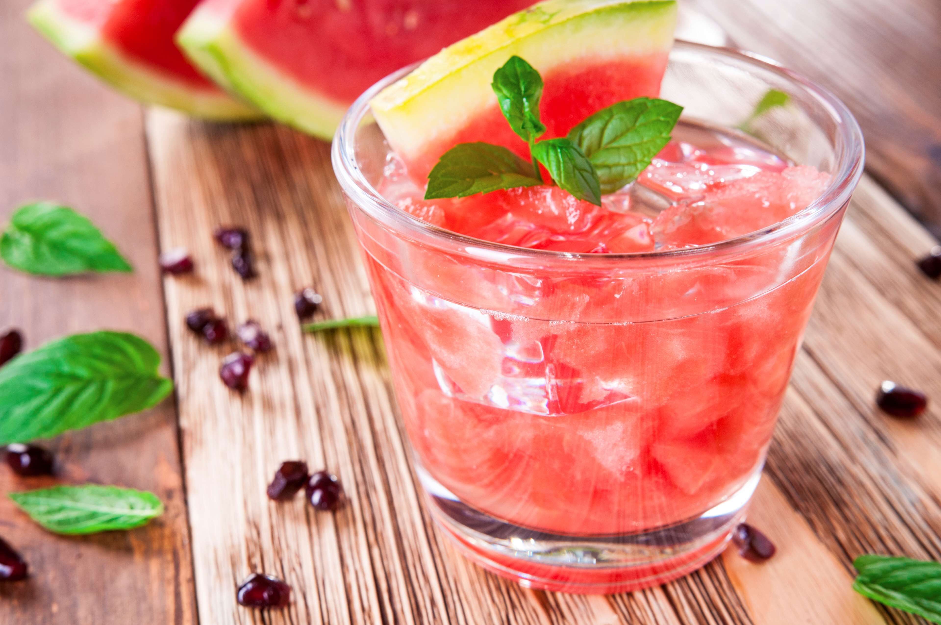 Un verre d’une boisson rose, servie avec des glaçons et décorée d’une tranche de fruit et d’une feuille de menthe, posé sur une surface en bois parsemée de baies et de feuilles.
