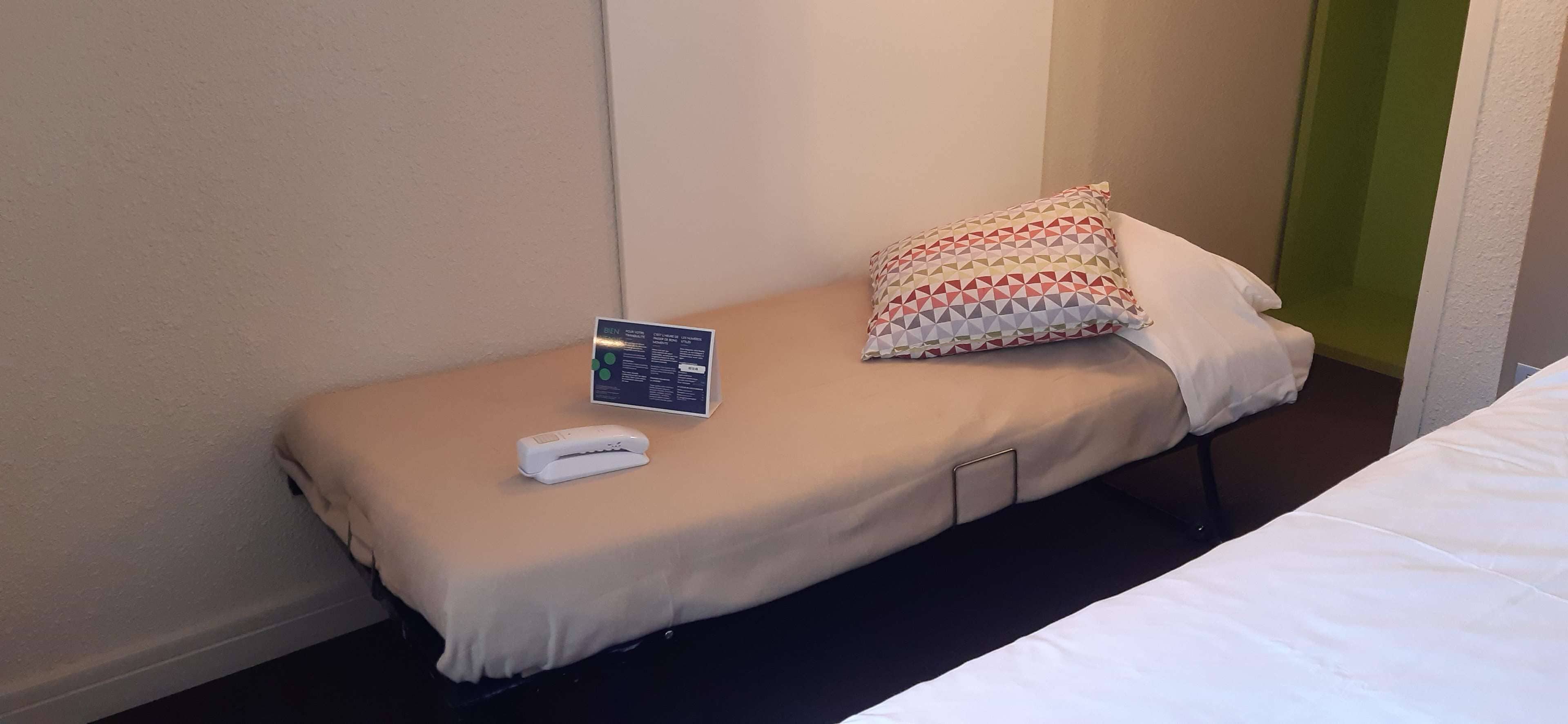 A single bed with a beige bedspread, a patterned pillow, a remote control, and a small informational card on the bed.