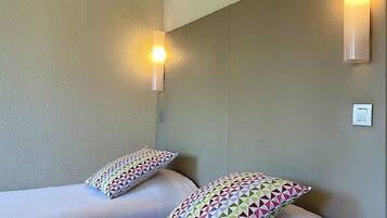 A hotel room with two beds, each with a colorful geometric-patterned pillow. There are wall-mounted lights on either side of the beds.