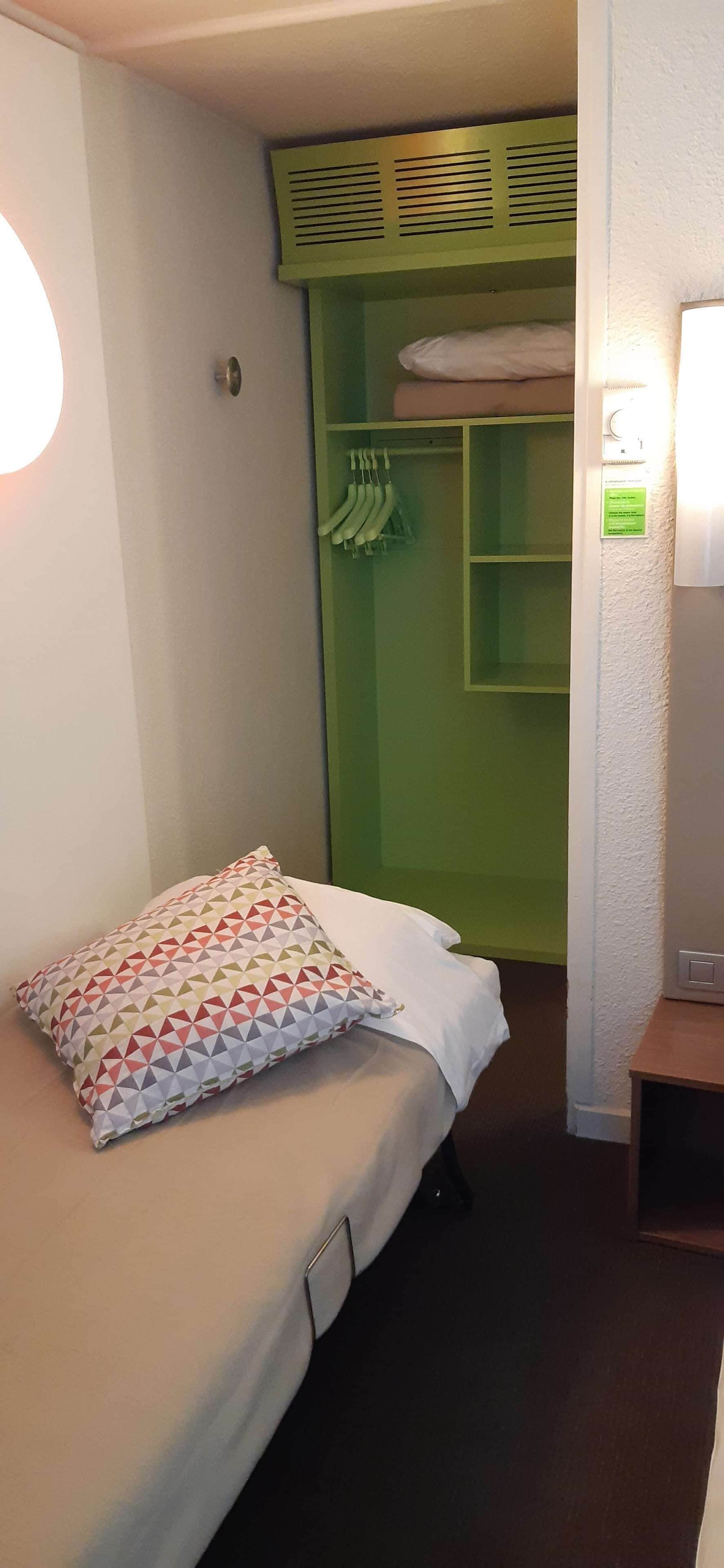 Un lit simple avec un oreiller à motifs, une armoire verte où sont suspendus des vêtements, et une lampe fixée au mur.