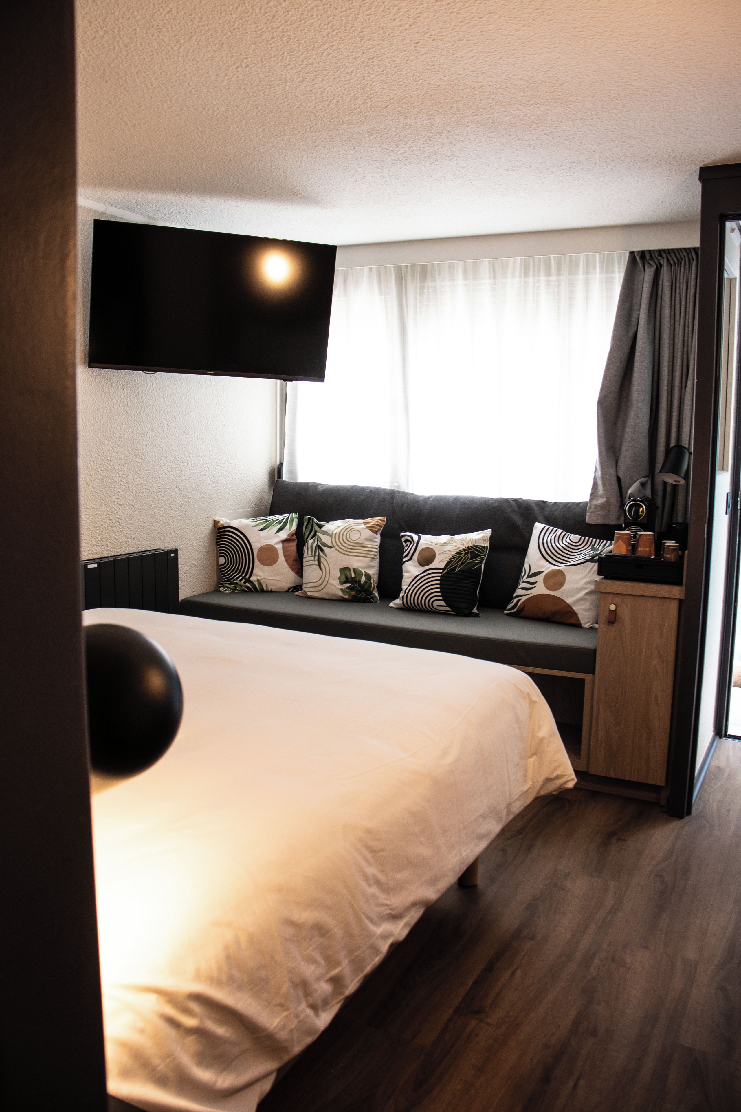 Habitación de hotel con una cama, un televisor de pantalla plana montado en la pared, una ventana con cortinas y una mesita de noche con una lámpara.