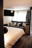 Hotellihuone, jossa on sänky, seinälle asennettu taulutelevisio, ikkuna verhoilla sekä y öpöytä lampulla.