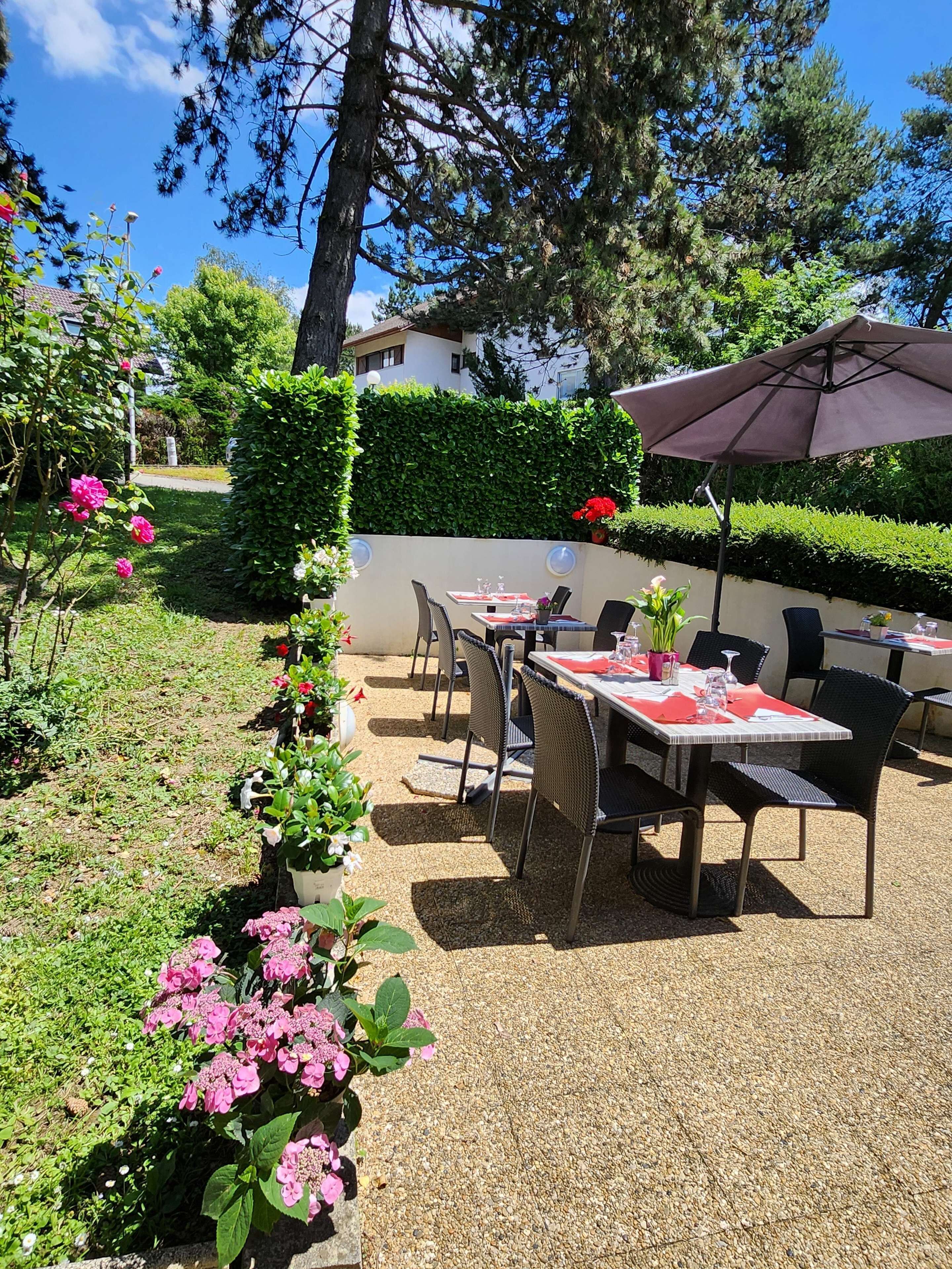 Un patio al aire libre con mesas y sillas, rodeado de vegetación y un seto.
