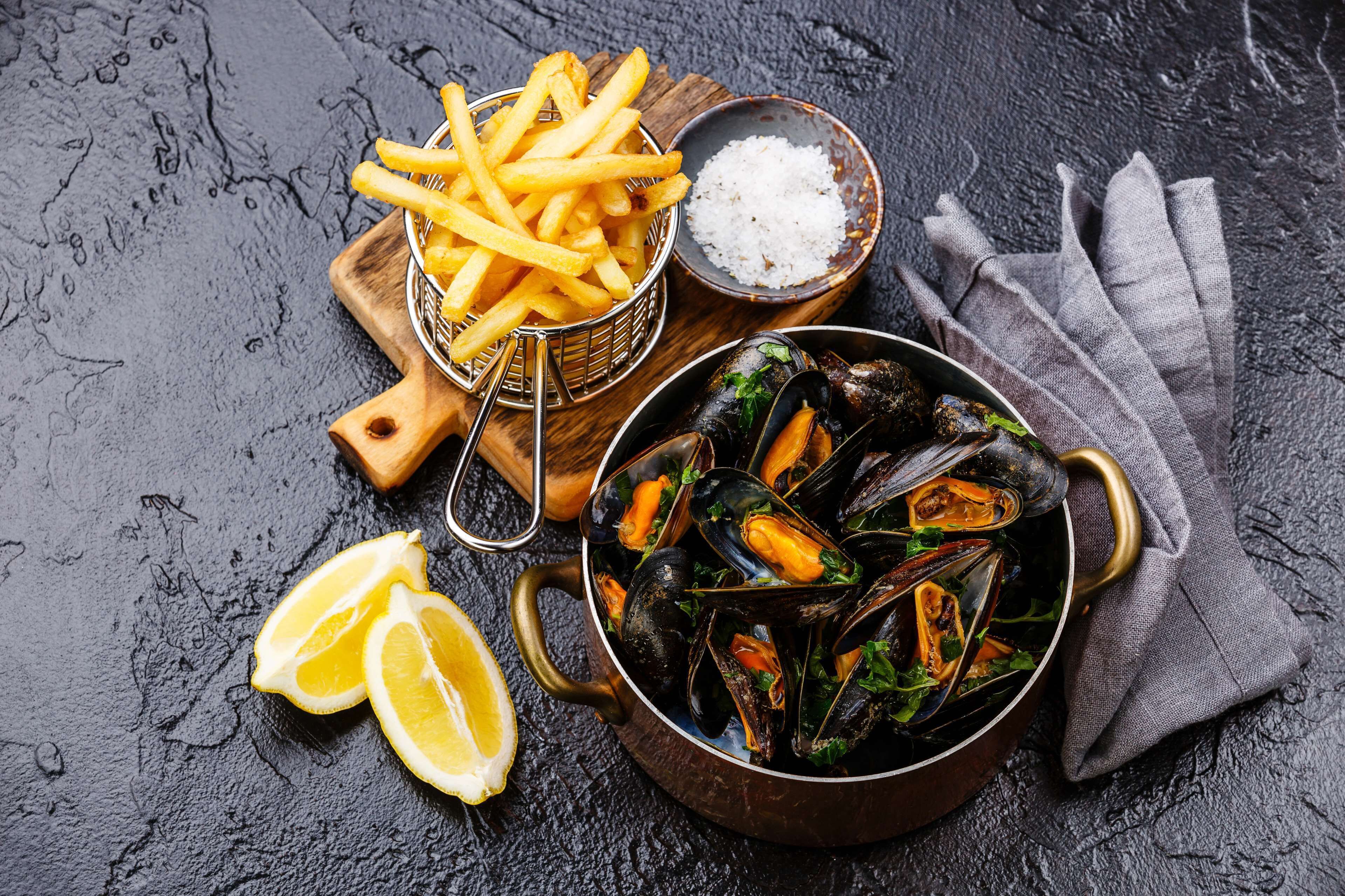 Mejillones en una olla, una cesta de papas fritas y un cuenco pequeño de sal sobre una superficie rugosa.