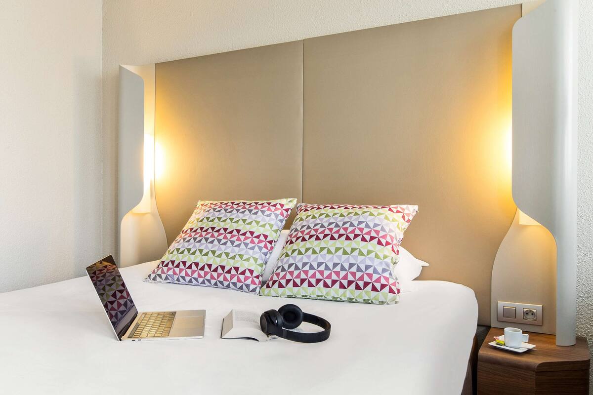 A neatly made bed with patterned pillows, a laptop, headphones, and a book on a white surface.