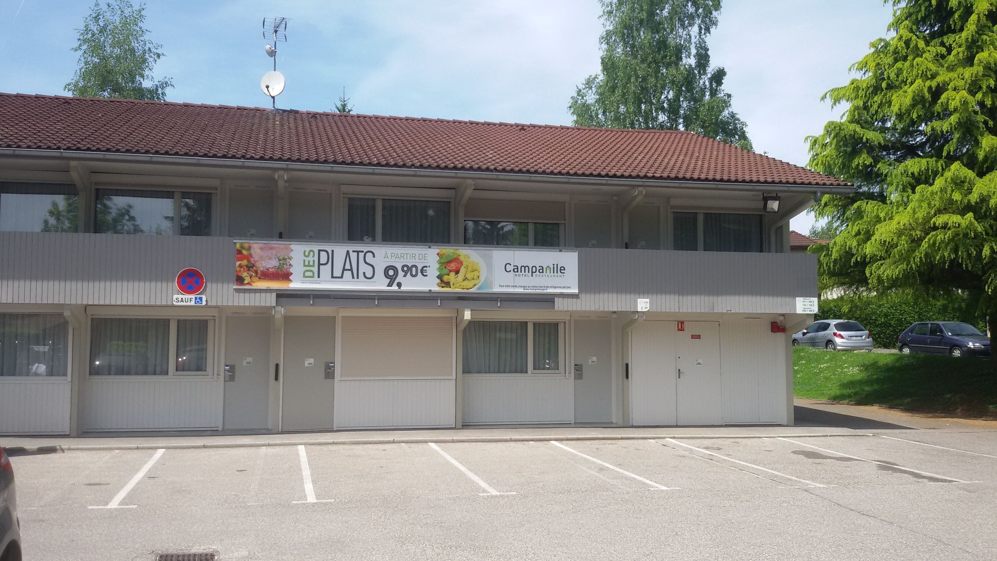 Un bâtiment à deux étages, avec un toit recouvert de tuiles rouges, une enseigne publicitaire pour un restaurant et un parking à l’avant.