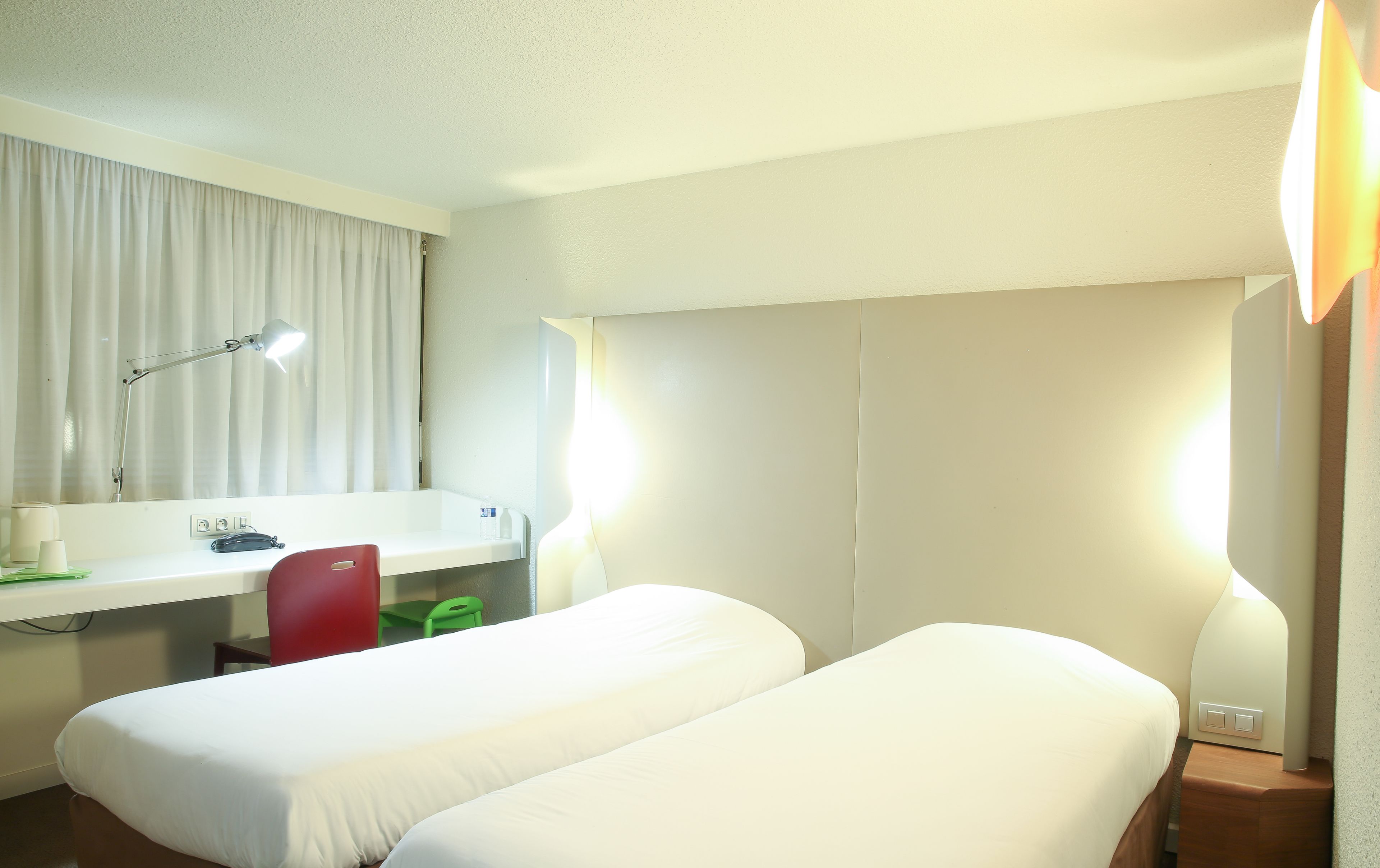 Una habitación de hotel con dos camas individuales, un escritorio con silla, un espejo, una lámpara y una ventana con cortinas.