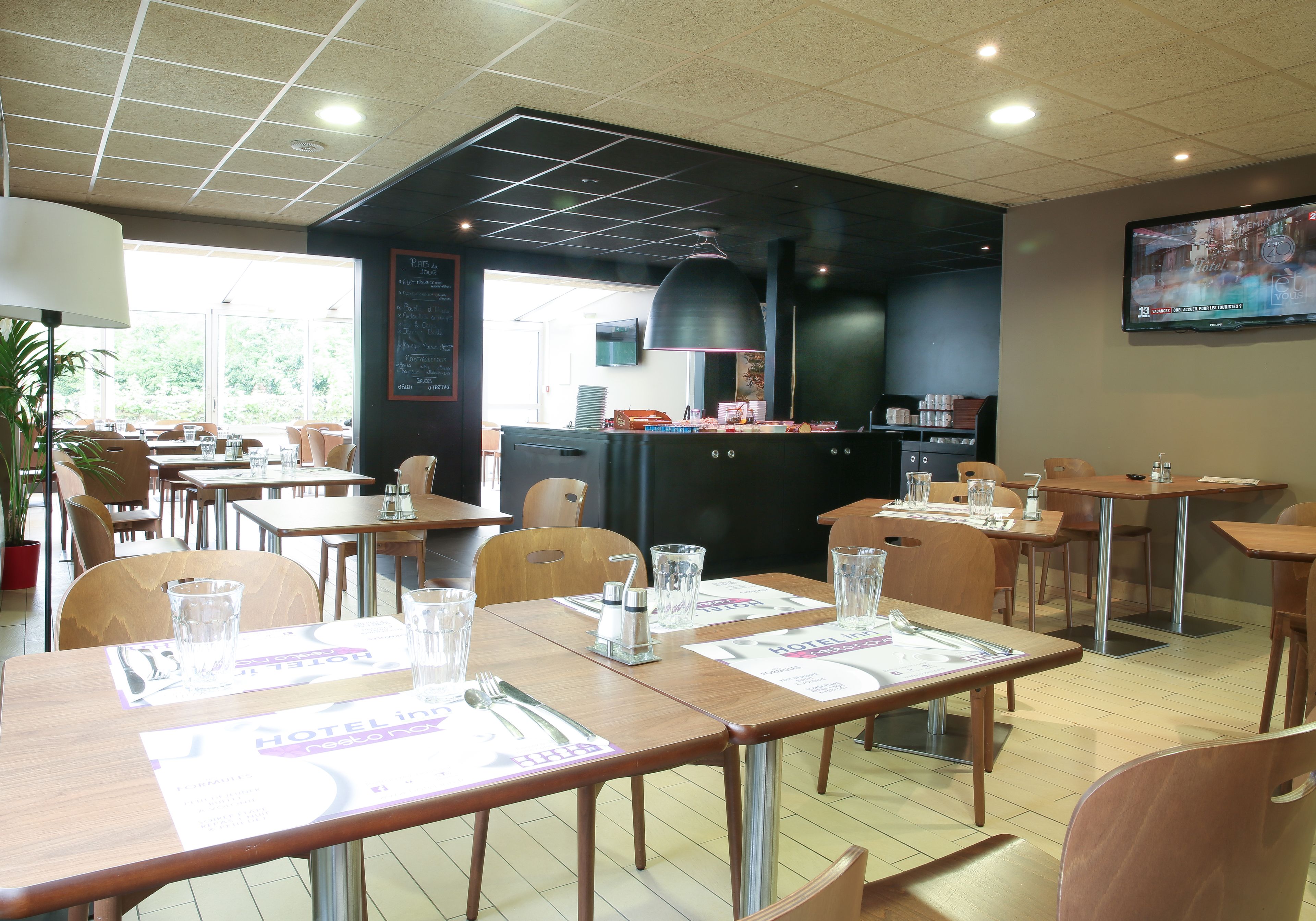 Un restaurante con mesas y sillas de madera, un amplio sector de cocina en color negro y un televisor fijado a la pared.