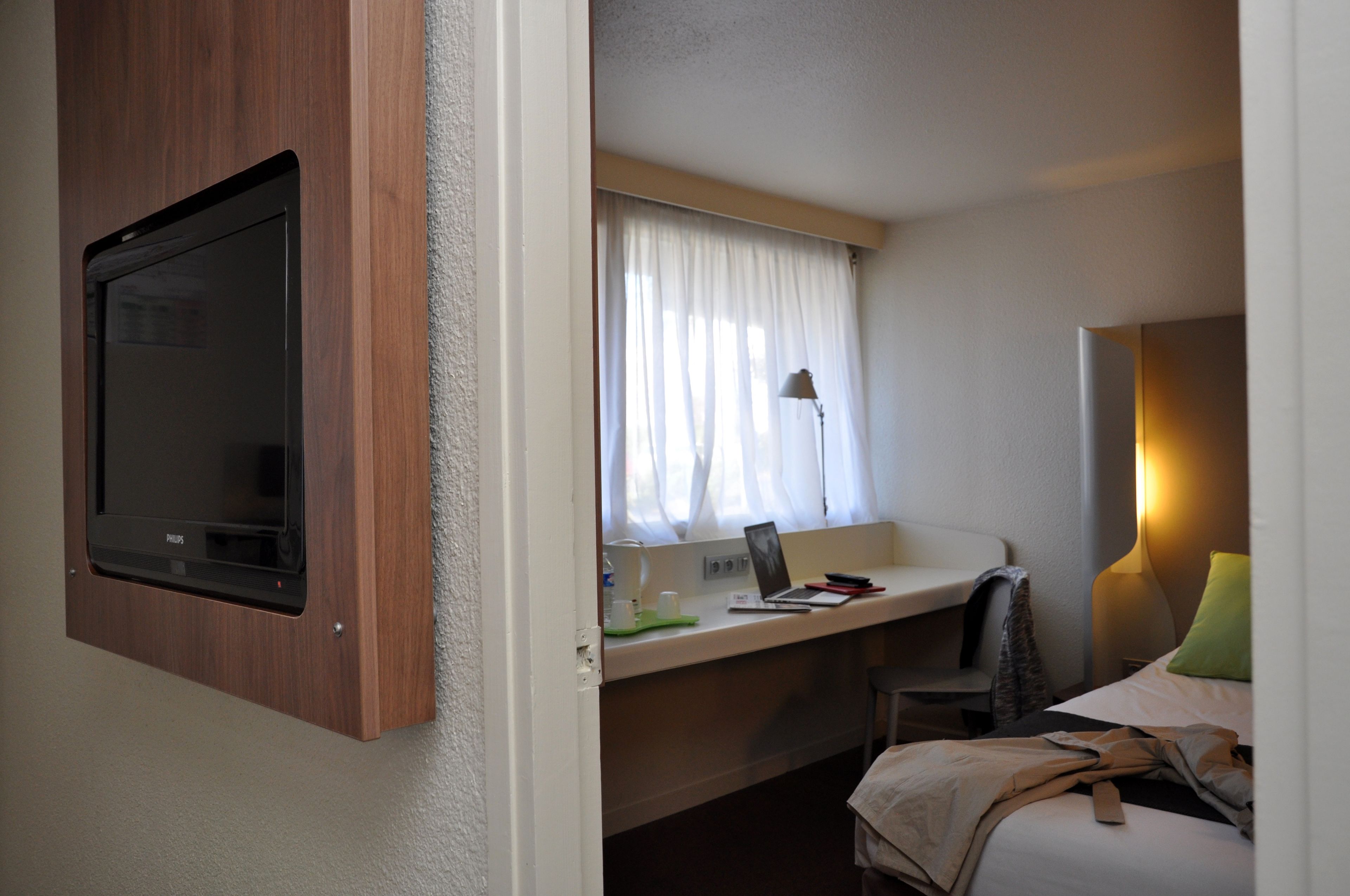 A hotel room with a built-in TV, a desk with a laptop, a lamp, and a bed with a pillow.
