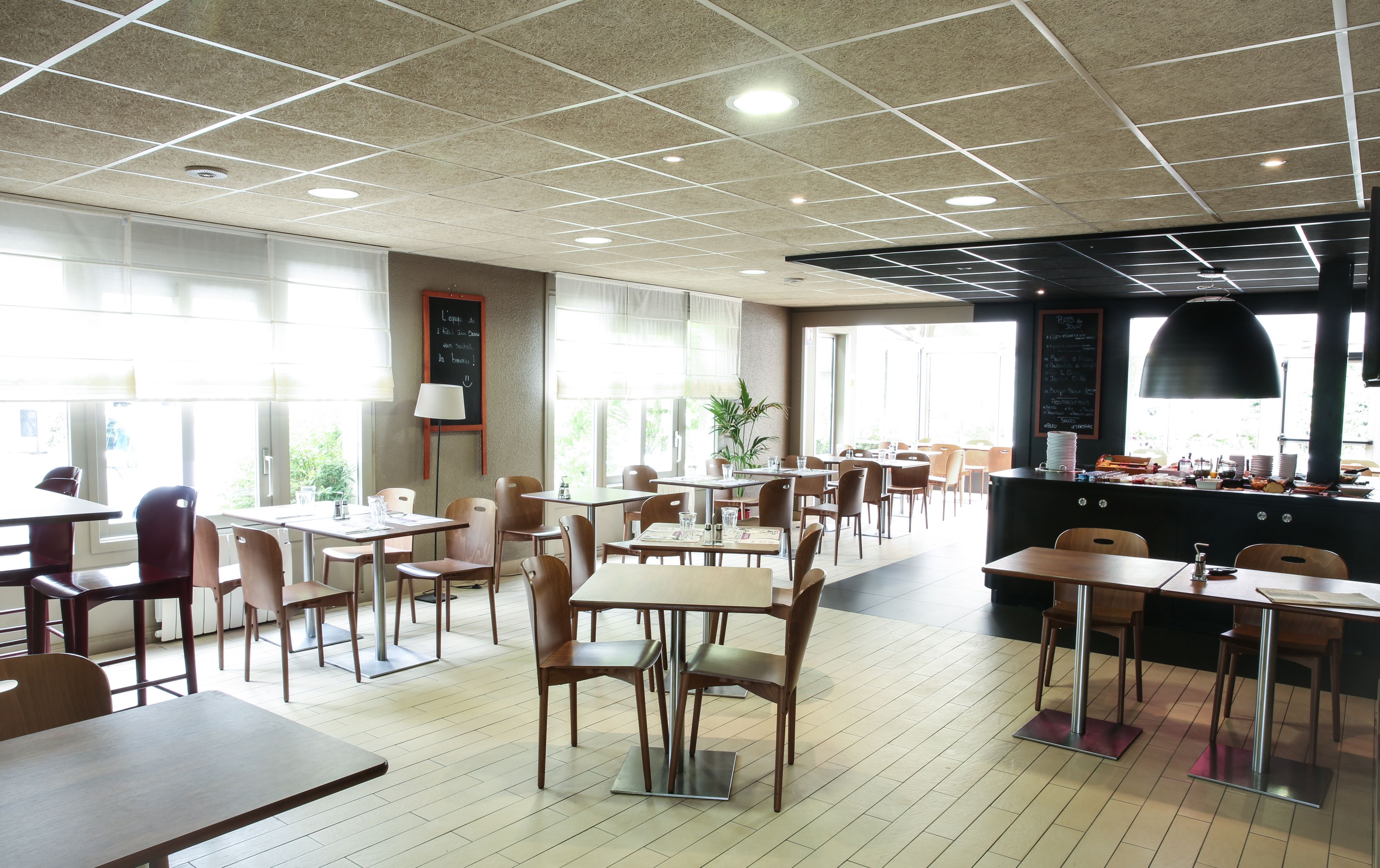 A restaurant with a high ceiling, grid-patterned ceiling, and natural light from large windows. Tables and chairs are arranged for dining.