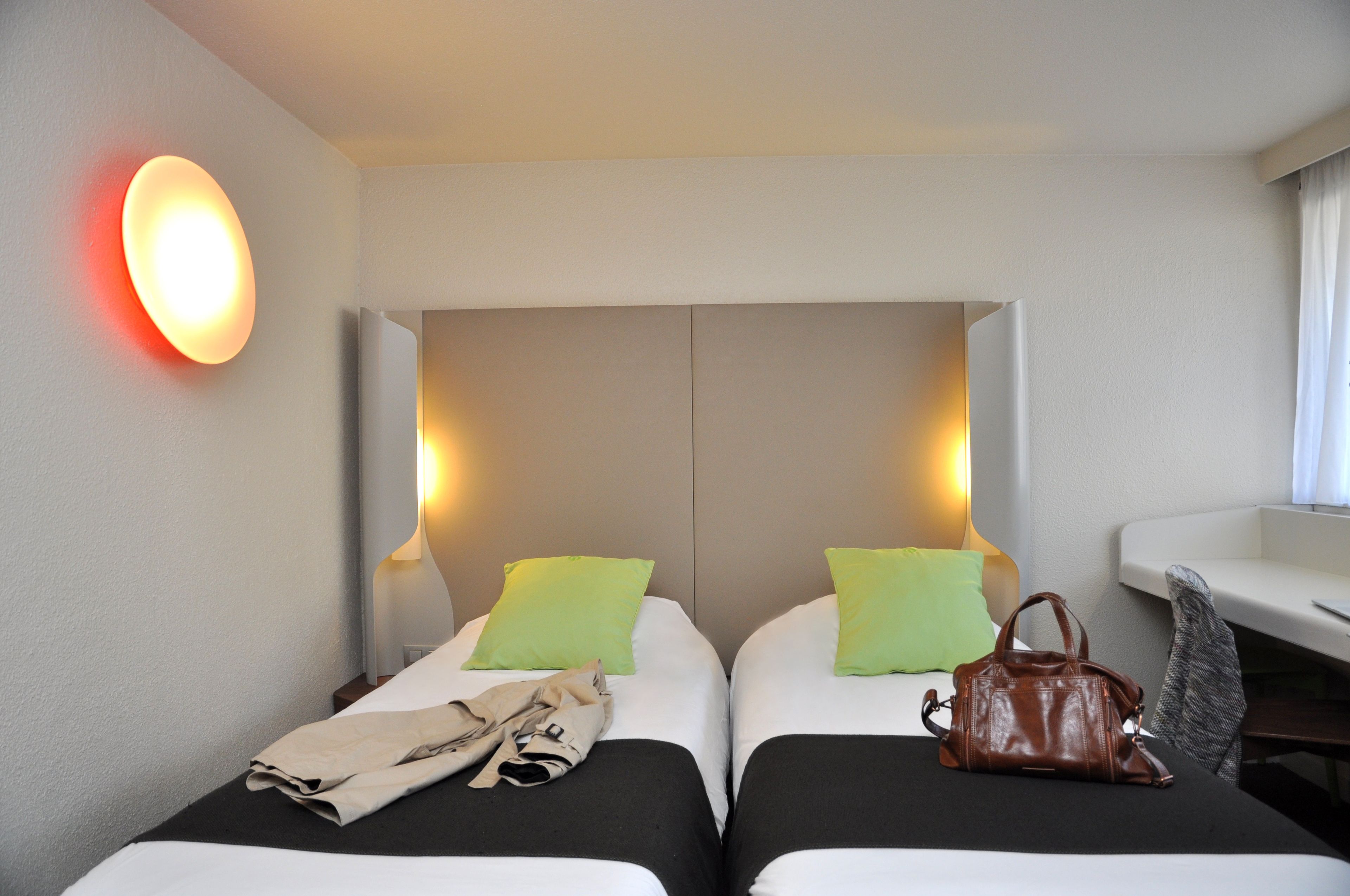Une chambre d’hôtel avec deux lits, une table de chevet, une lampe, un sac à main et une fenêtre avec des rideaux.