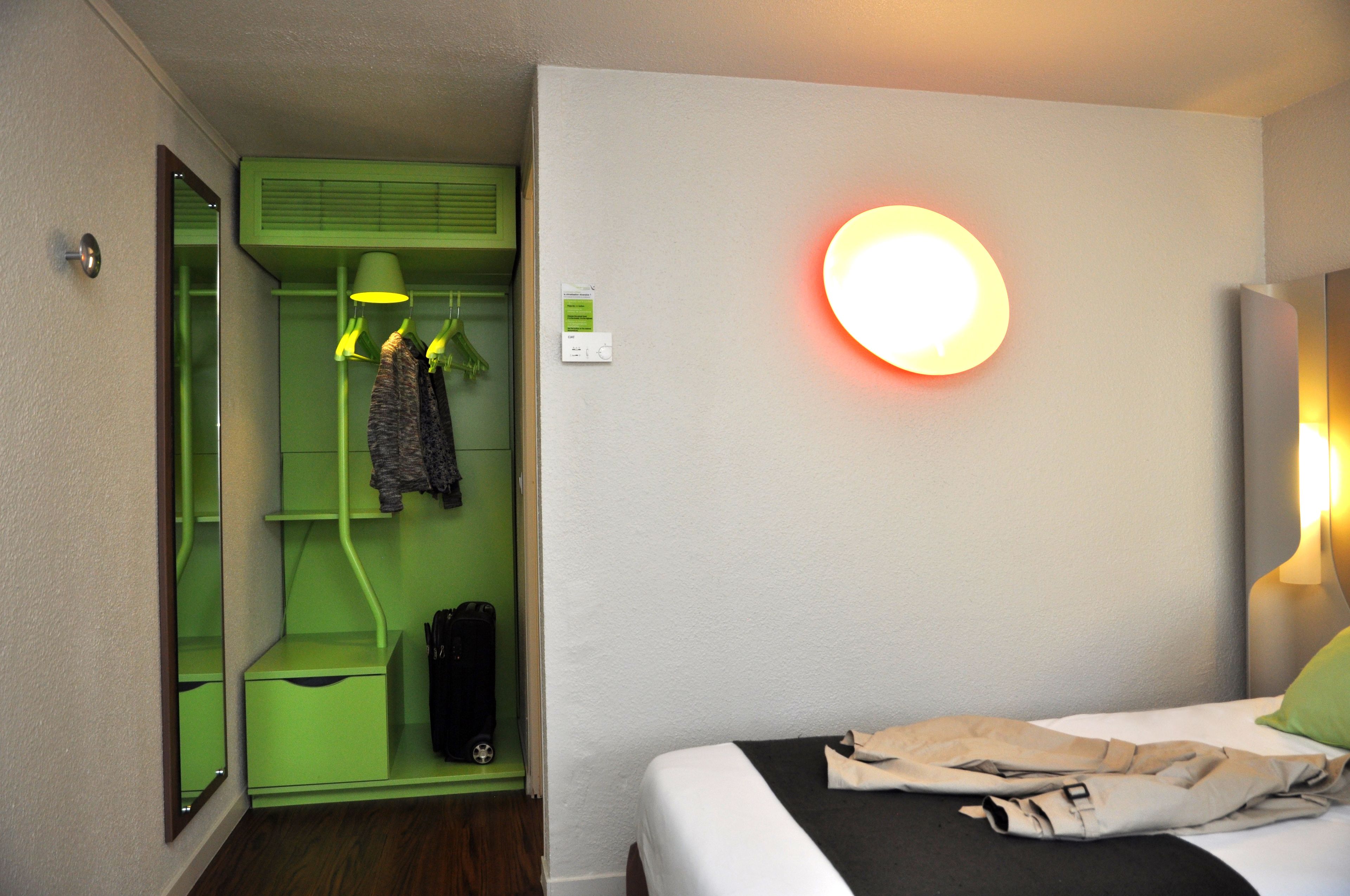 Une chambre d’hôtel avec un lit, une armoire verte, un miroir et une lampe fixée au mur.