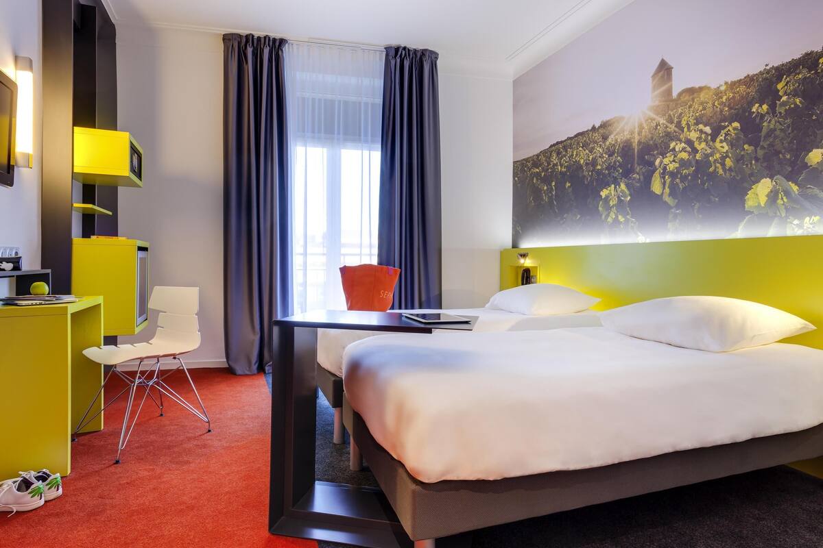 Habitación de hotel con cama, escritorio, silla, televisión y un amplio ventanal con cortinas.