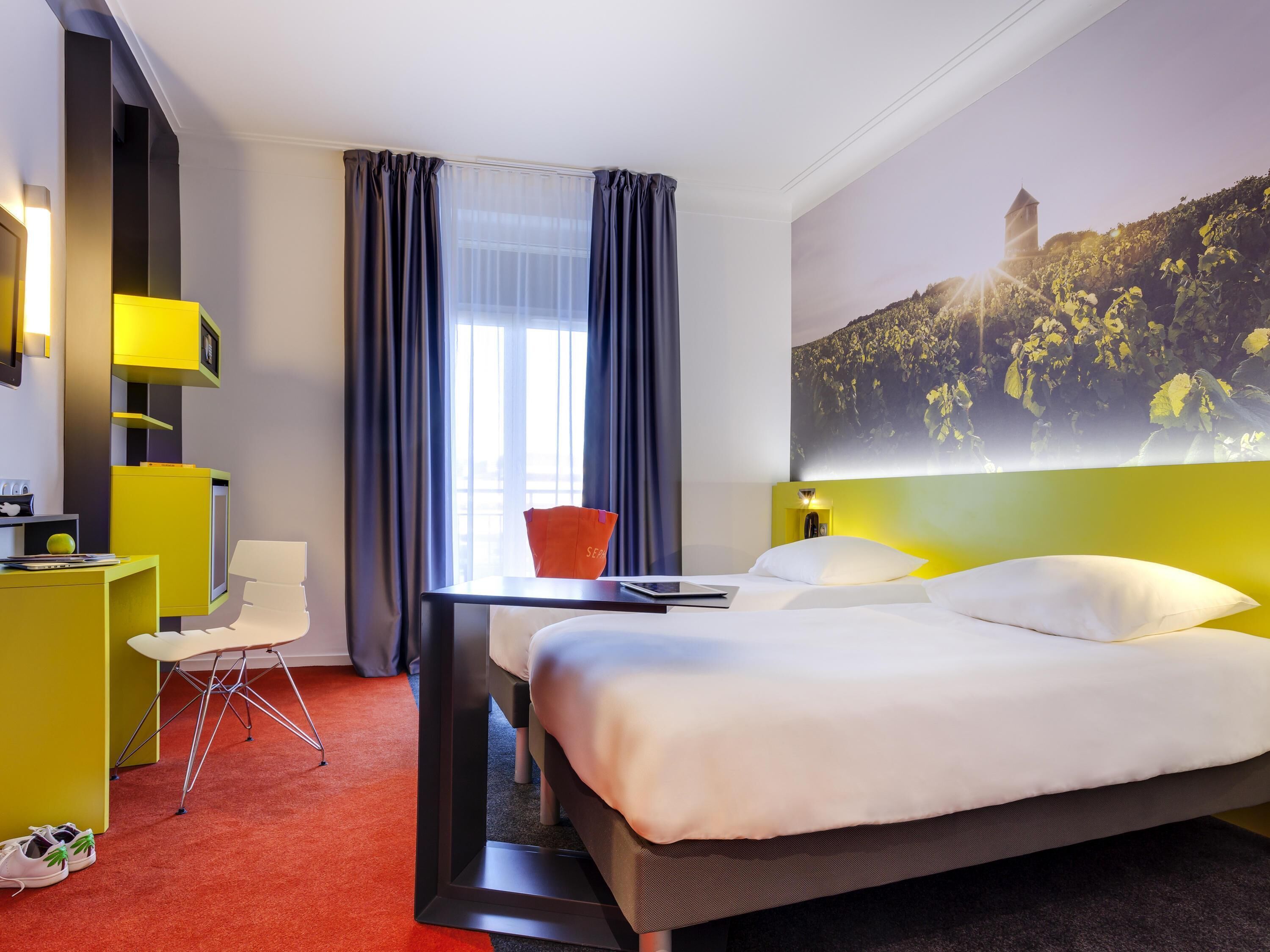 Habitación de hotel con cama, escritorio, silla, televisión y un amplio ventanal con cortinas.