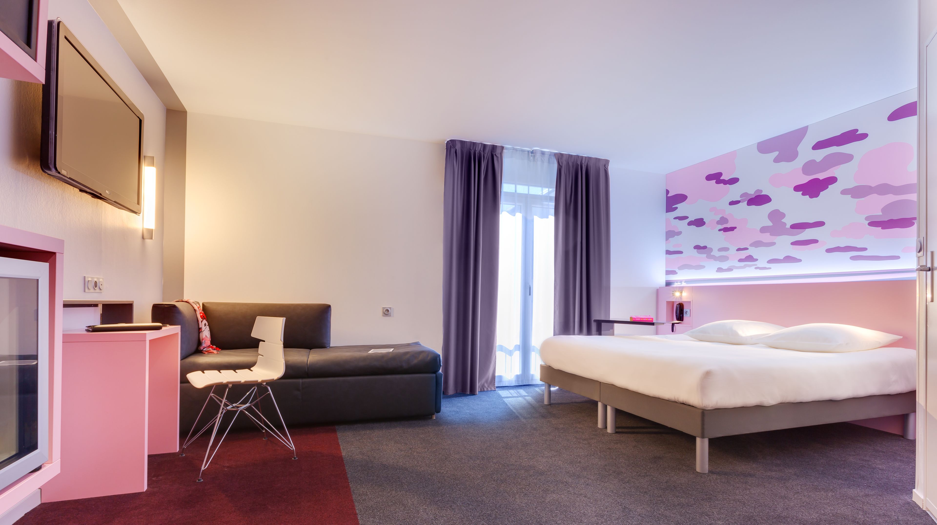A modern hotel room with a bed, a desk, a chair, and a television.