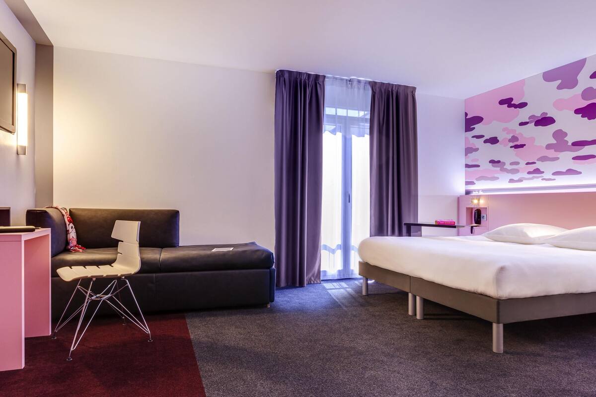 Quarto de hotel com cama, sofá, escrivaninha e uma janela grande com cortinas.