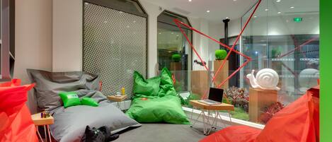 Habitación de hotel moderna con cama, cojines verdes y rojos, un ordenador portátil sobre una mesita auxiliar y una pared de cristal con vistas a la ciudad.