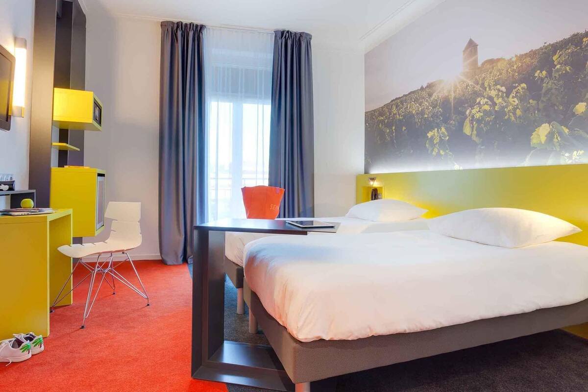Habitación de hotel con cama, escritorio, silla, ventana con cortinas y lámpara de pared.