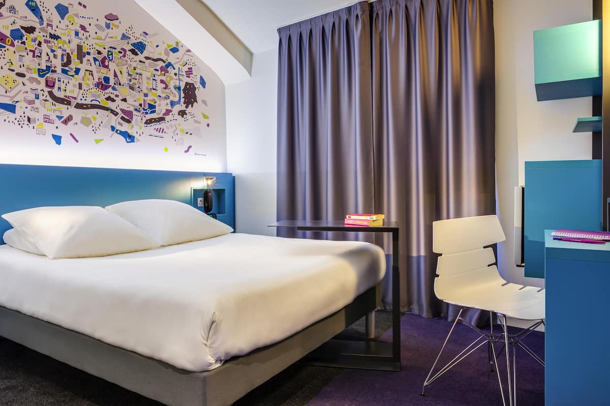 A modern hotel room with a large bed, a desk, a chair, a wall art piece, and a curtain.
