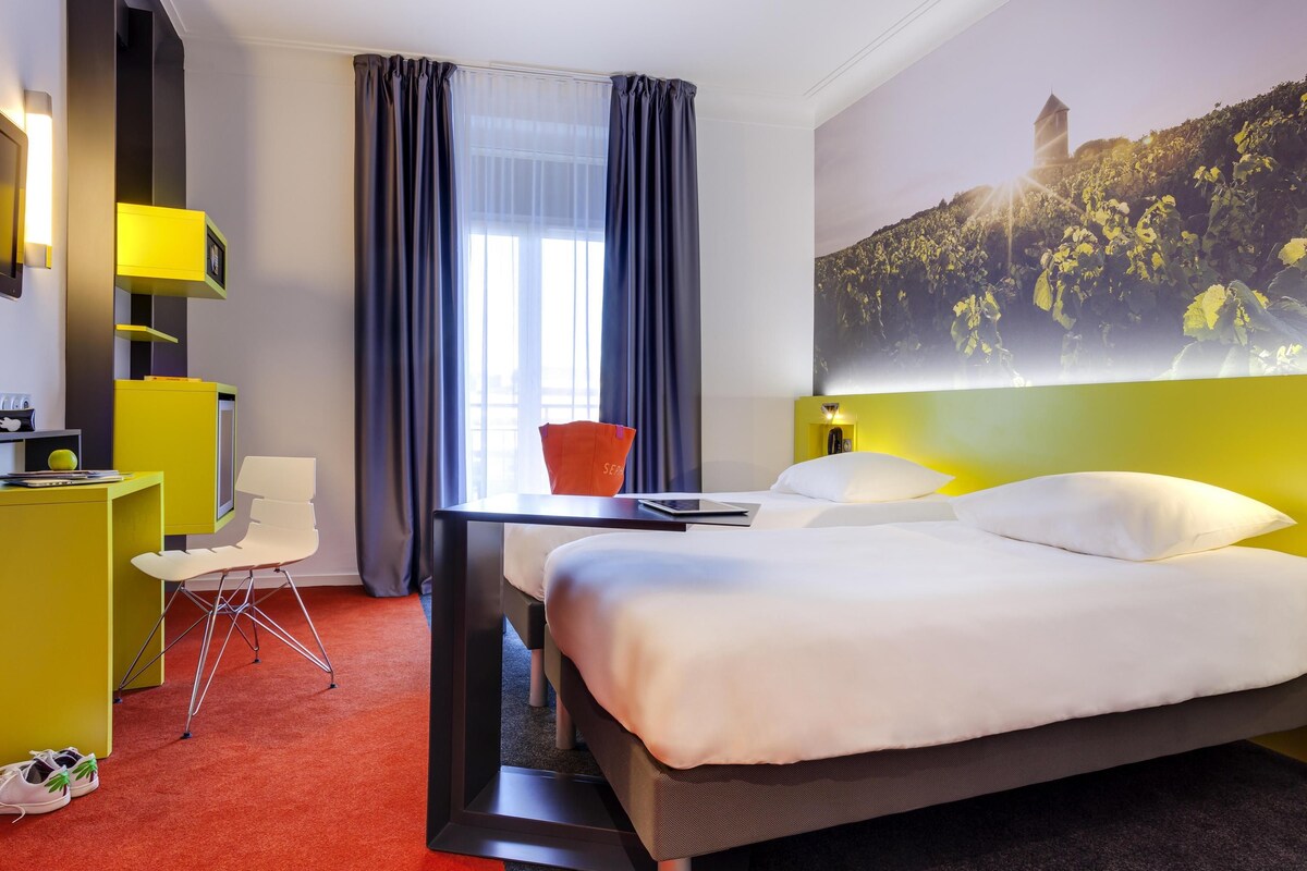 Habitación de hotel con cama, escritorio, silla, televisión y un amplio ventanal con cortinas.