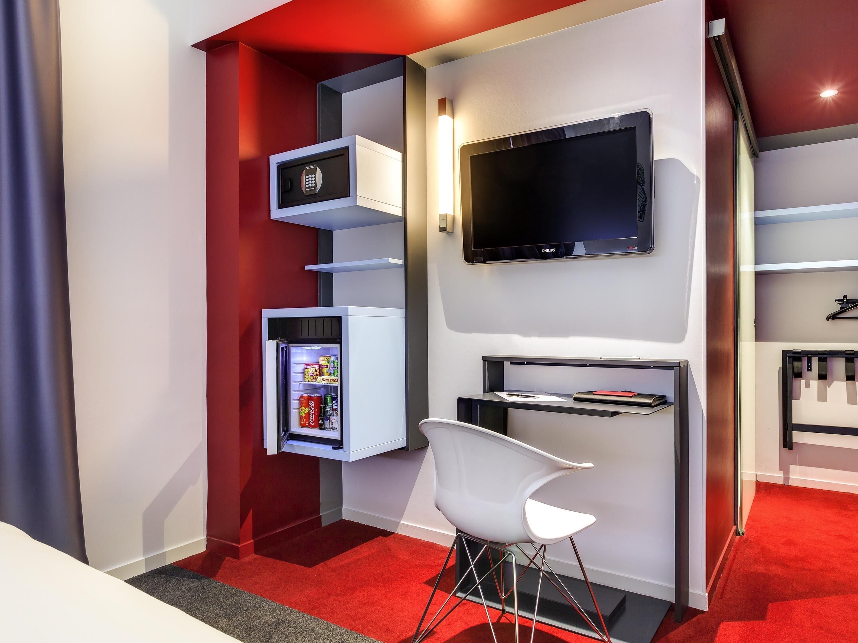 A modern hotel room with a built-in mini-fridge, a flat-screen TV, a white chair, and a red carpeted floor.