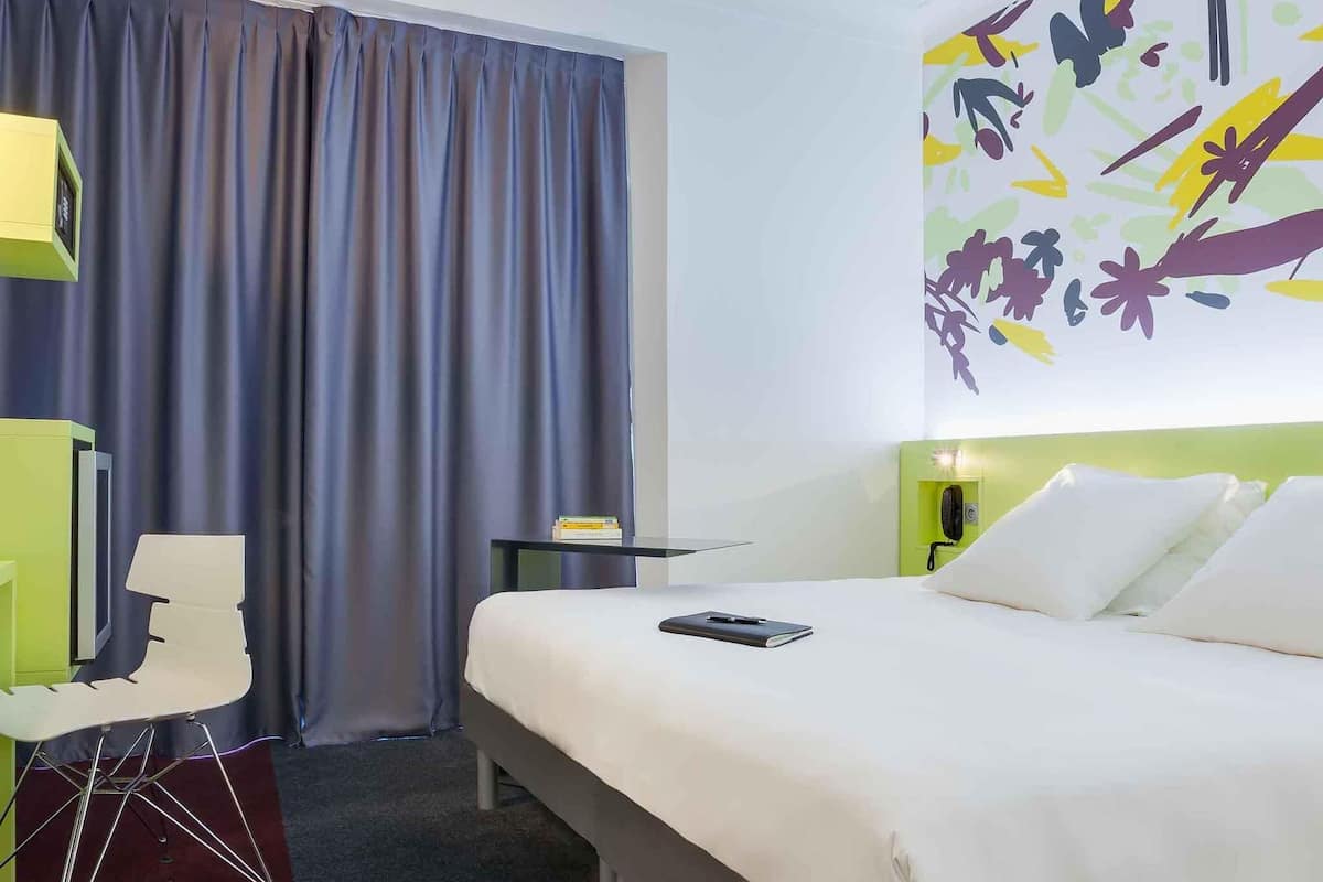 Una habitación de hotel con cama, escritorio, silla, cortina y una pared decorada con arte abstracto.