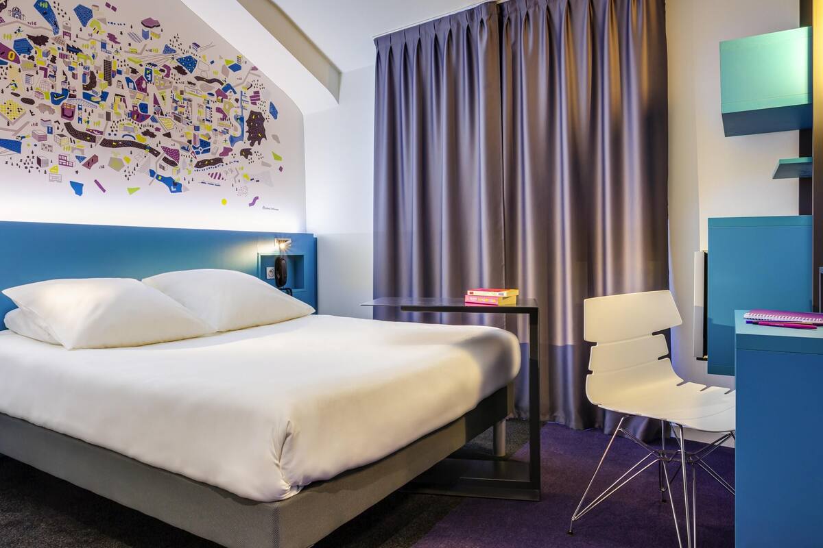 Quarto de hotel moderno com uma cama grande, uma escrivaninha, uma cadeira, uma obra de arte na parede e uma cortina.